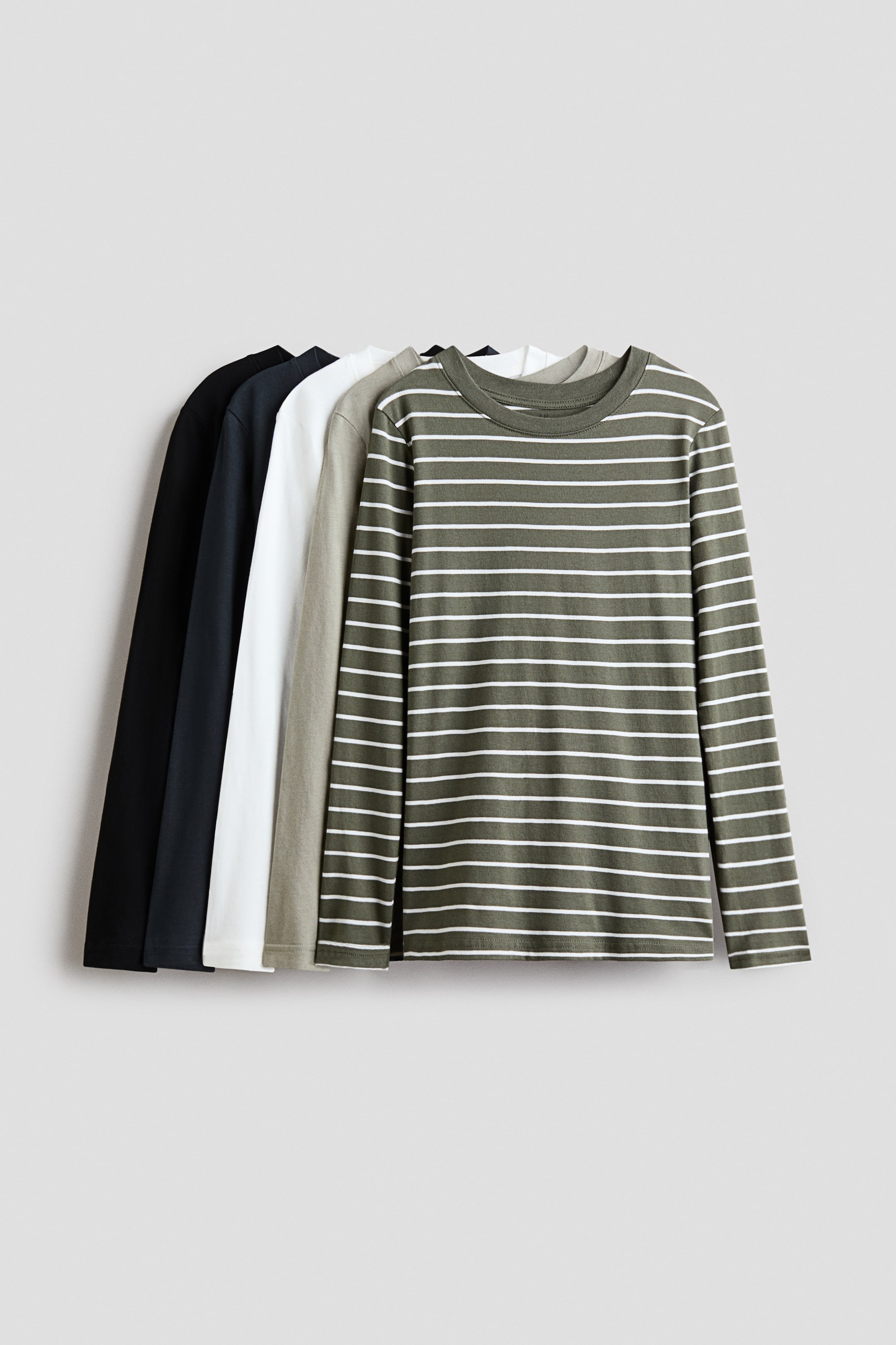 Agrandir l'image: Cinq t-shirts en jersey de coton à manches longues sont suspendus dans une composition frontale superposée. Le t-shirt placé au premier plan présente un motif à rayures horizontales vert olive et blanches, avec un col rond bordé de côtes, tandis que les t-shirts unis disposés derrière lui apparaissent en gris clair, blanc, bleu marine foncé et noir.