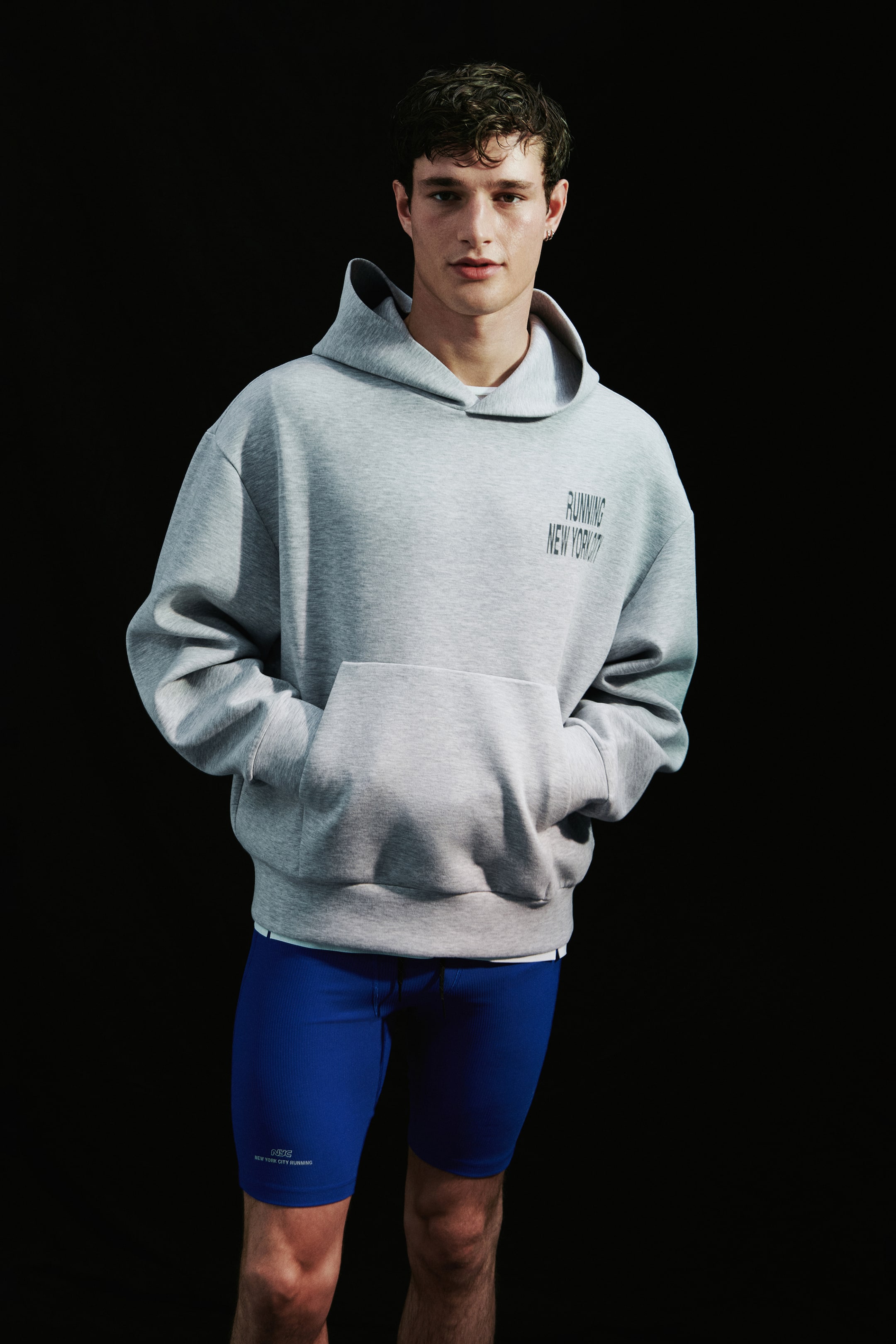 View larger image: A light gray oversized hoodie featuring a front kangaroo pocket and black graphic text on the left chest is being worn by the model, who is placing both hands in the pocket. The hoodie is styled with bright blue, form-fitting athletic shorts.