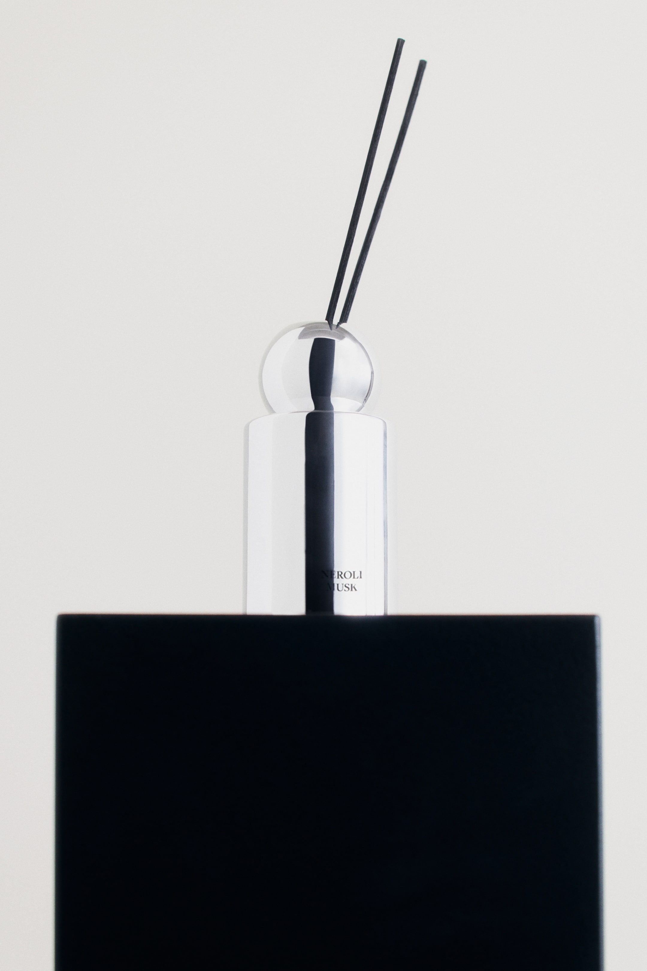 Vis større bilde: En sylinderformet duftpinner med en reflekterende kromfinish står på en solid svart blokk, med teksten "NEROLI MUSK" på forsiden. To slanke, svarte duftpinner strekker seg oppover fra en gjennomsiktig, sfærisk glasskork på toppen.