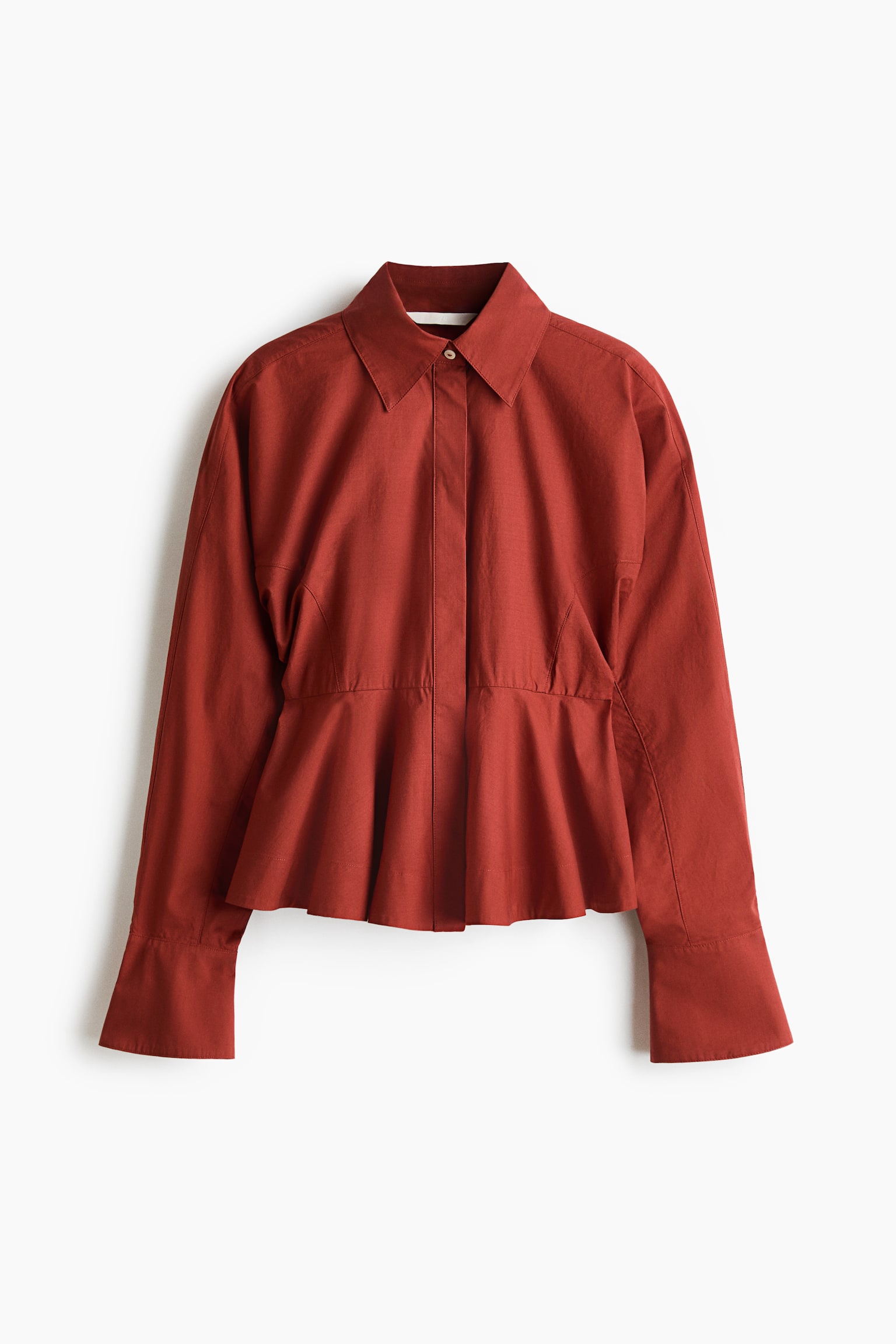 A rust-red cotton poplin shirt is displayed facing front, featuring a pointed collar, a concealed button placket, and long batwing sleeves with wide, flared cuffs. The garment is gathered at the waist, flaring out into a structured peplum hem.