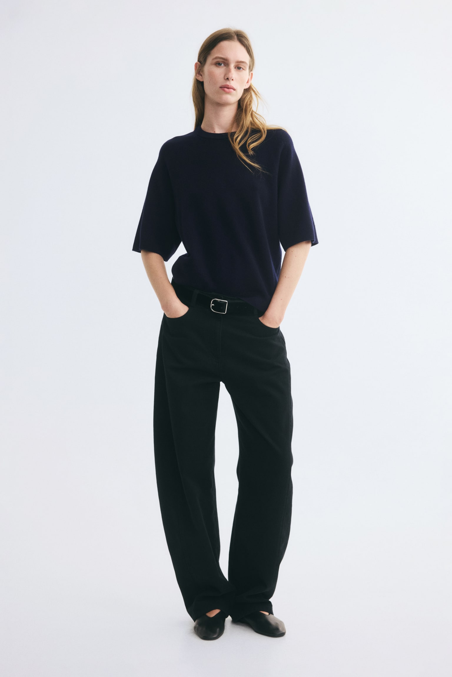 The model is wearing a dark navy knitted wool top featuring a round, rib-trimmed neckline and elbow-length raglan sleeves. The top is styled tucked into black wide-leg trousers secured with a black belt. - 1