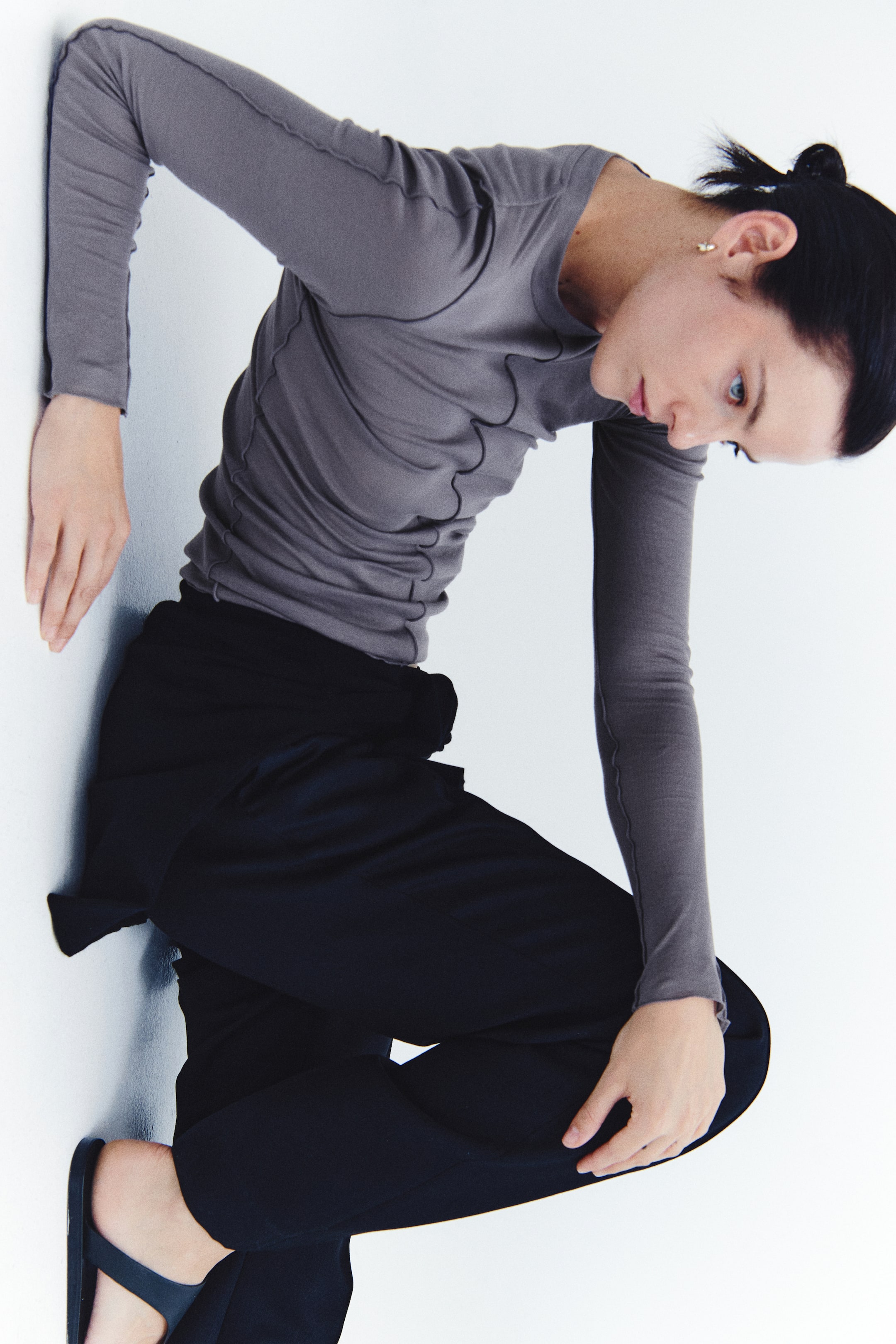 View larger image: A grey long-sleeved top featuring wavy joined seams and a round neckline is being worn by the model, who is posing in a leaning position. The top is styled with black trousers.