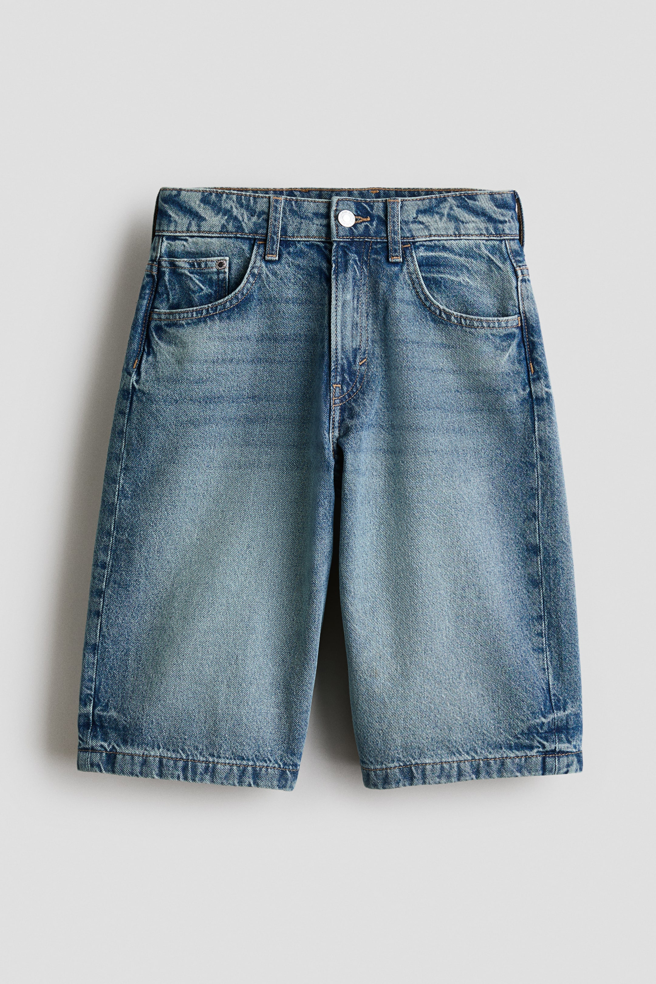 Se større billede: Knælange shorts i vasket bomuldsdenim præsenterer en frontvisning, med to forlommer, en møntlomme, bæltestropper og en lynlås med knaplukning. De viser en løs pasform gennem hofte og lår med en sænket skridtsøm.
