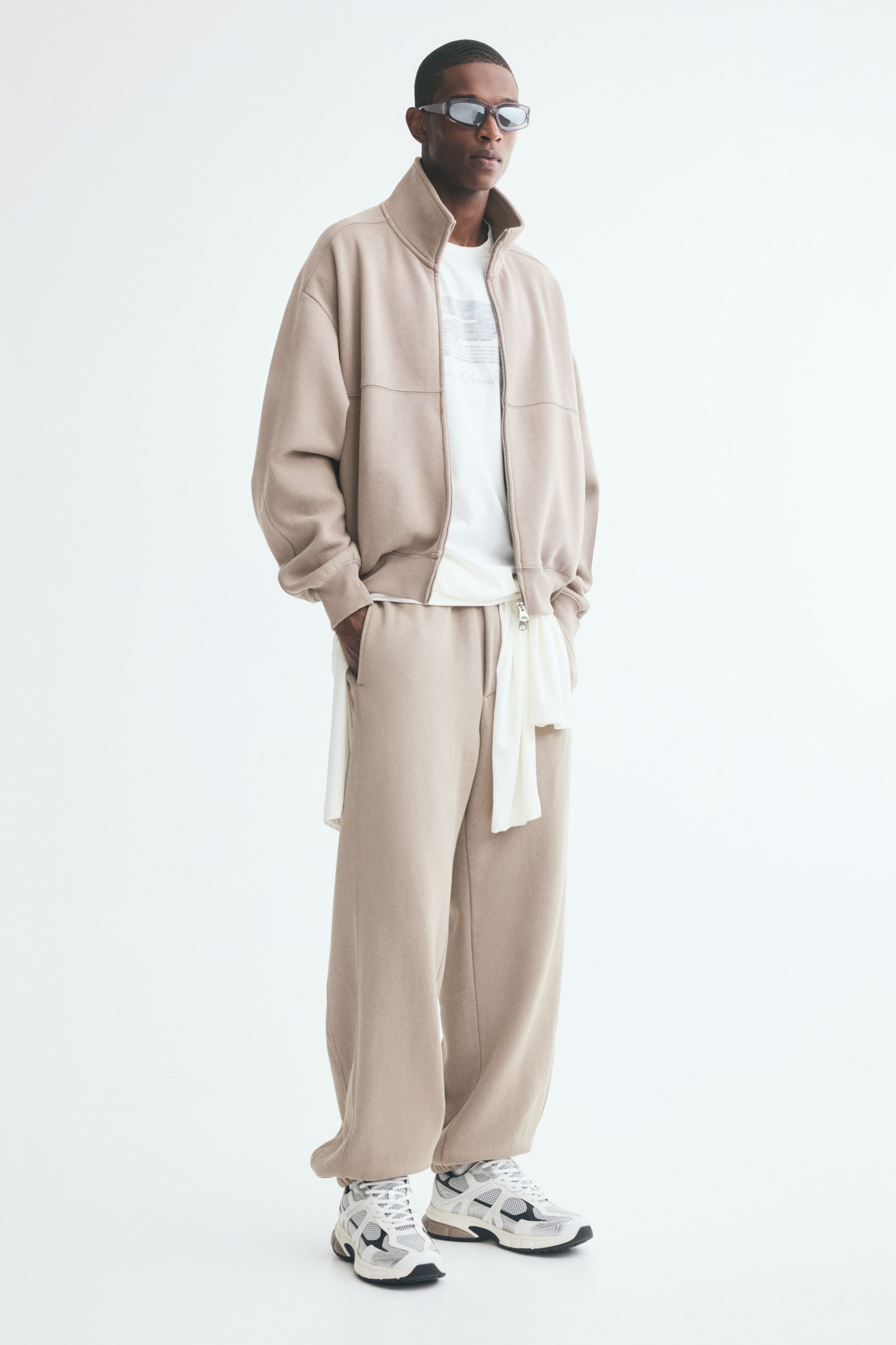 Voir une image plus grande: Pantalon de survêtement beige à coupe décontractée, avec jambes droites, coutures avant visibles et ourlets élastiqués, porté par le mannequin. Le mannequin est debout, une main glissée dans une poche latérale, et porte le pantalon avec un blouson zippé assorti, un T-shirt blanc et des chaussures de sport.