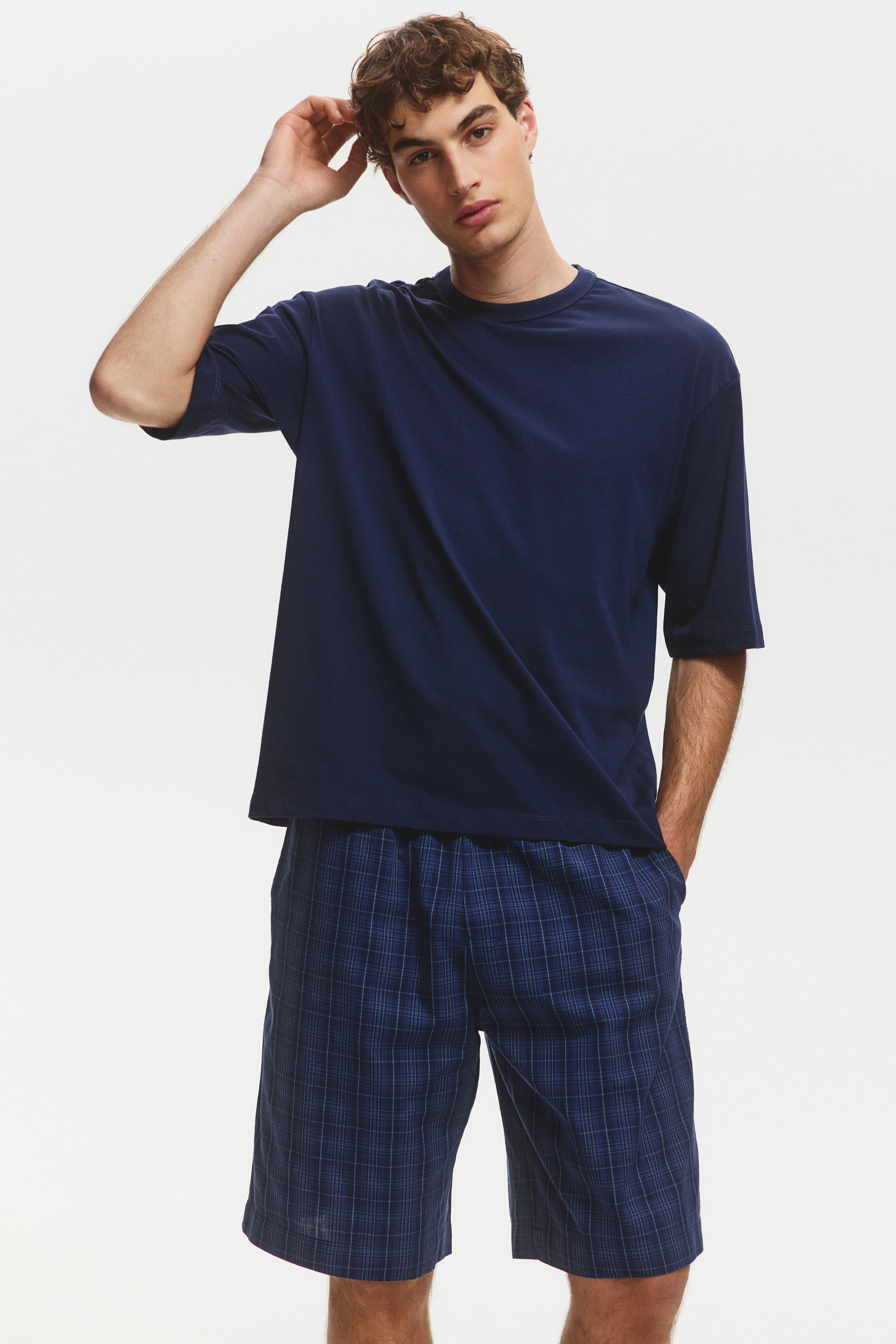 拡大画像を表示: ネイビーブルーのクルーネックでリラックスフィットの半袖Tシャツが、ブルーのチェック柄ショーツと組み合わせられています。ショーツは、ウエスト部分にゴムが入っており、リラックスしたカットで、膝の少し上までの長さです。