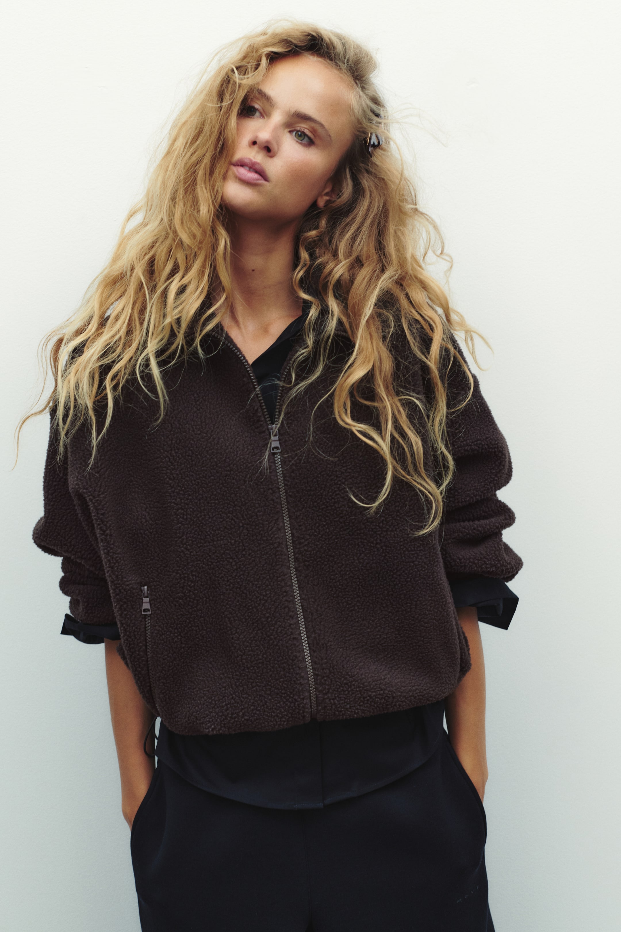 View larger image: A dark brown, loose-fit teddy jacket featuring a stand collar, dropped shoulders, and a front zip is worn by the model. The jacket, which includes a zipped side pocket, is layered over a black collared shirt and black trousers.