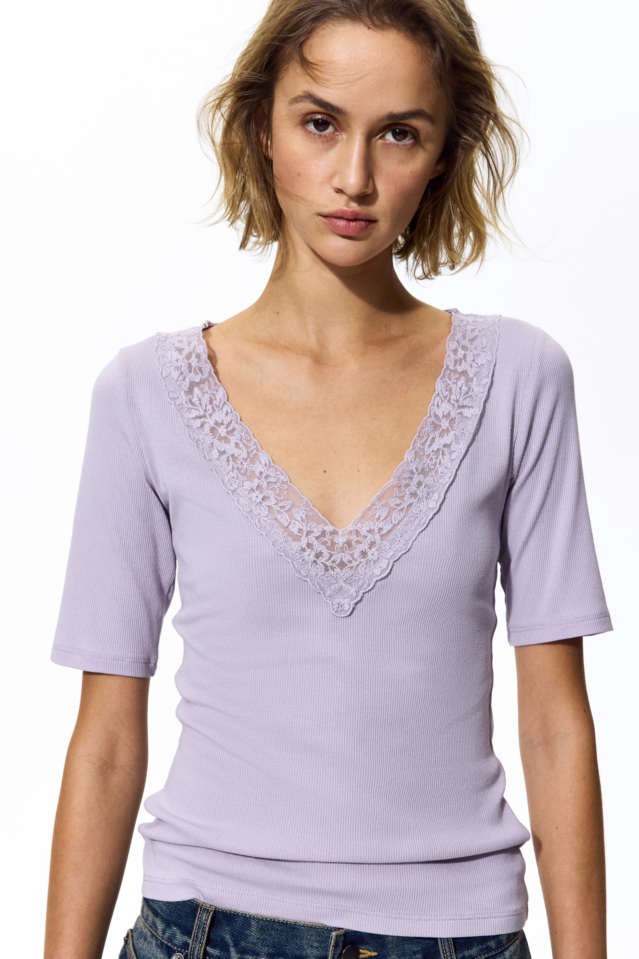 Büyük resmi görüntüle: Açık mor renkli, nervürlü triko üst, şeffaf çiçek desenli dantel süslemeli V yaka ve kısa kollu olarak sergileniyor. Giysi, vücuda oturan bir siluet sunuyor.