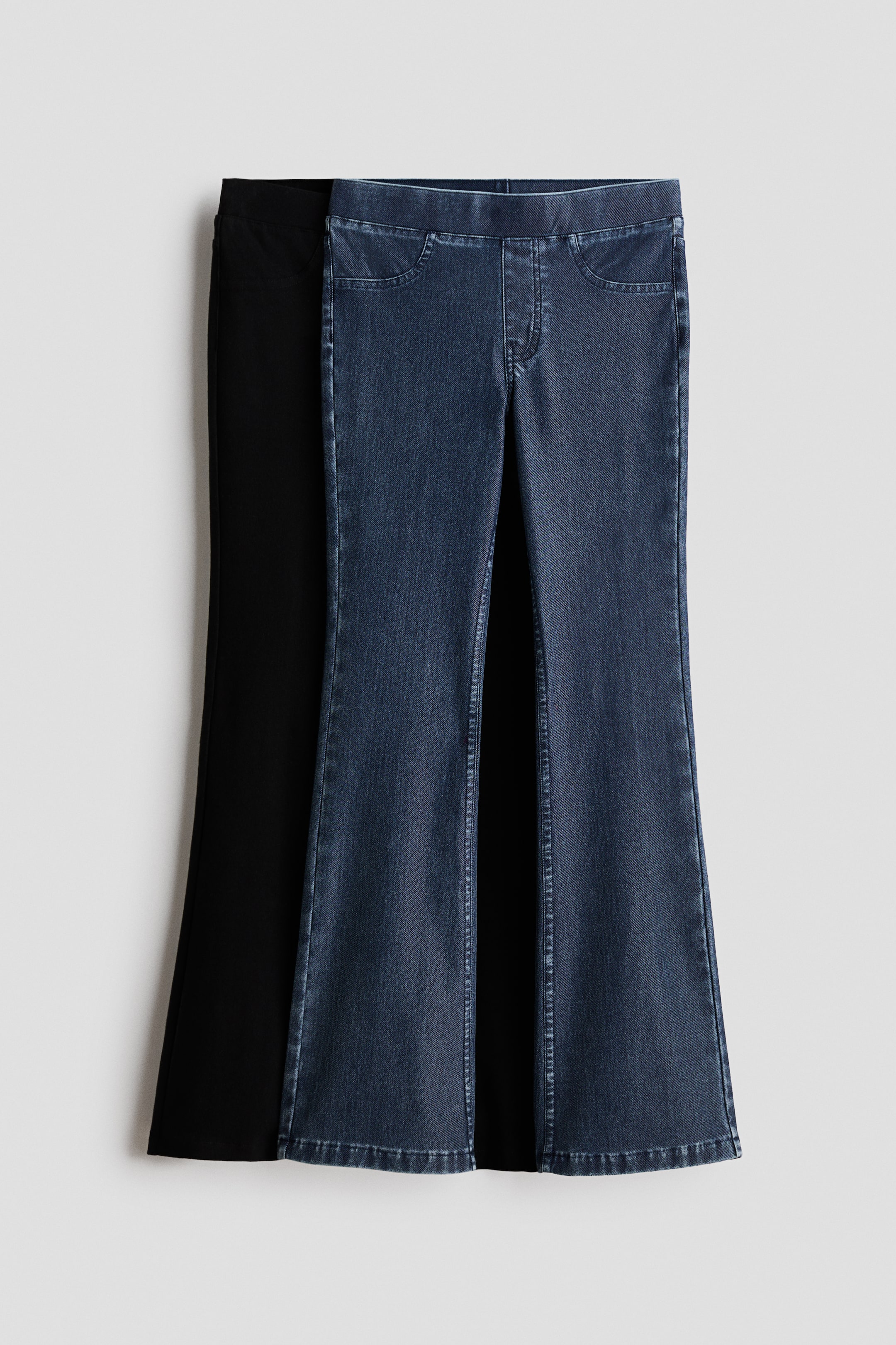 View larger image: Two pairs of flare pants are on display, facing forward. The front pair showcases a dark blue denim-like fabric, a pull-on waistband, faux front pockets, and a faux fly, while a solid black pair is partially visible behind it, also styled in a flare design.
