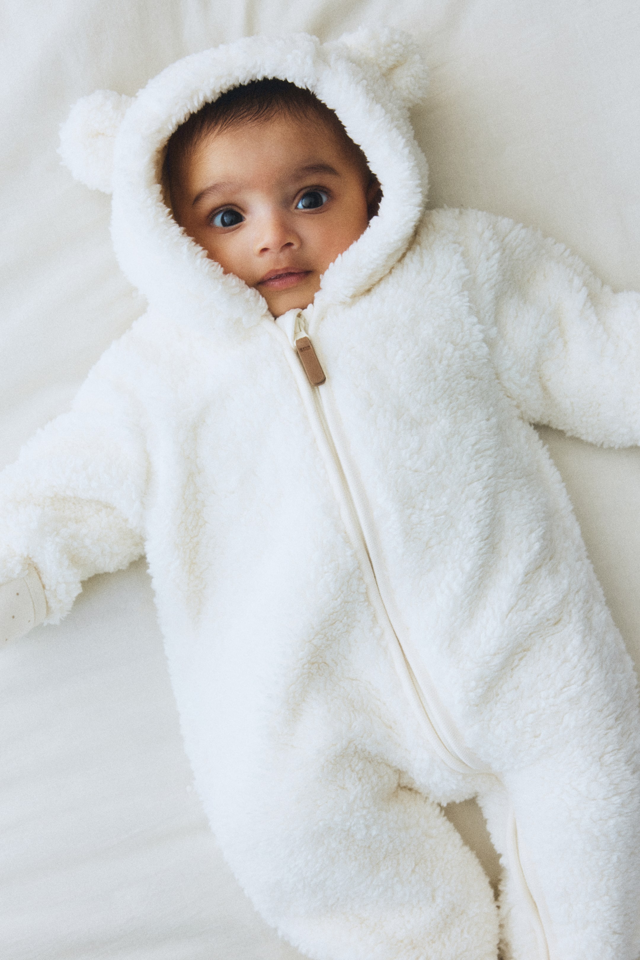 Vis større bilde: En hvit, lodden baby-body vises fra et fugleperspektiv, med en hettegenser med bjørneører og en hel lengde frontglidelås med en brun trekkflik. De lange ermene viser en hvitprikket innvendig mansjett.
