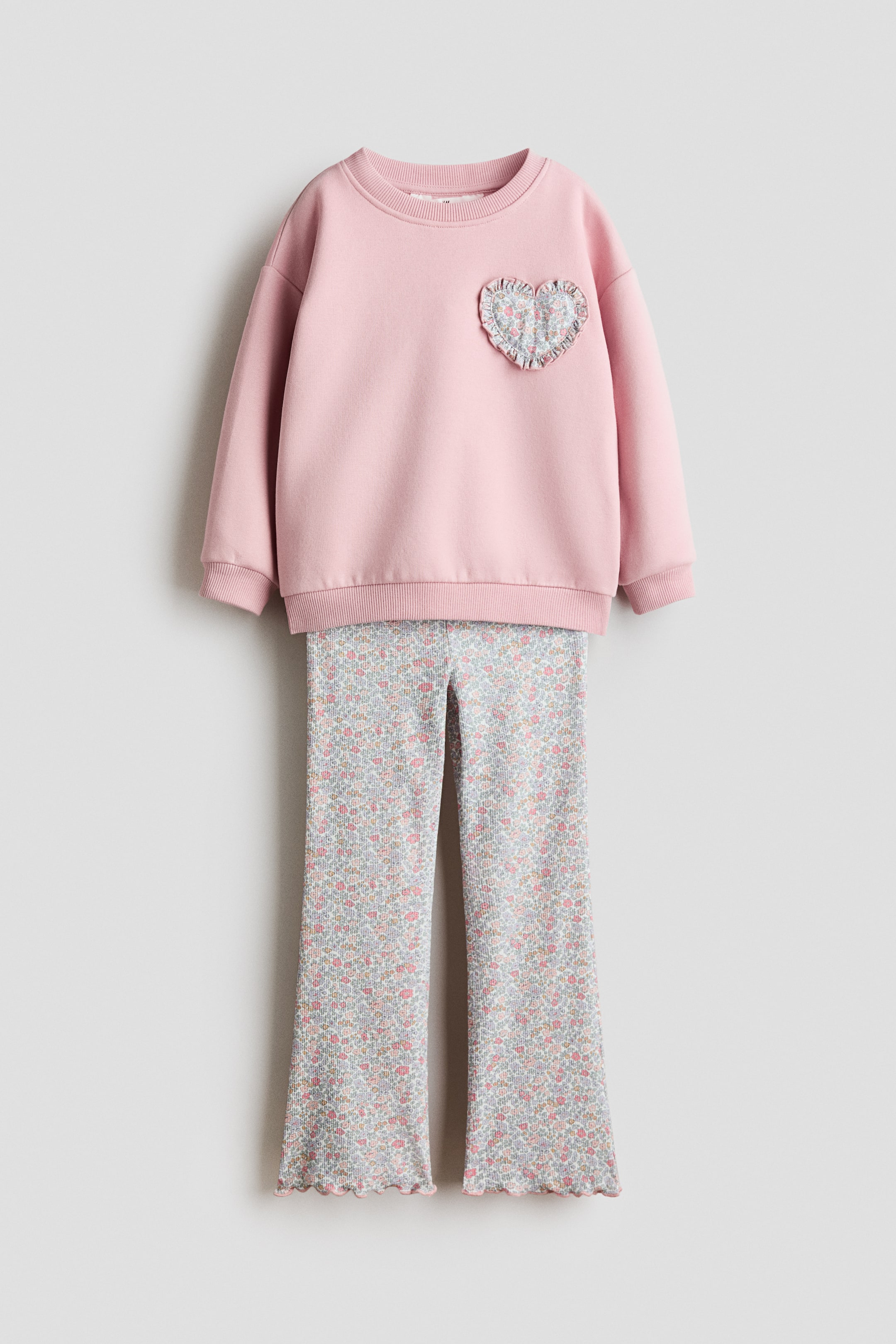 View larger image: A 2-piece sweatshirt and leggings set is showcased, featuring a pink crew-neck sweatshirt with ribbed cuffs, a ribbed hem, and a ruffled heart-shaped patch adorned with a floral pattern on the left chest. The coordinating ribbed leggings display an all-over floral print and lettuce-edge hems.