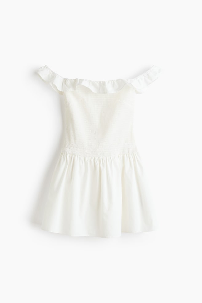 A white off-the-shoulder dress is displaying a ruffled neckline, a smocked bodice, and a gathered waist flowing into a flared skirt. - 1