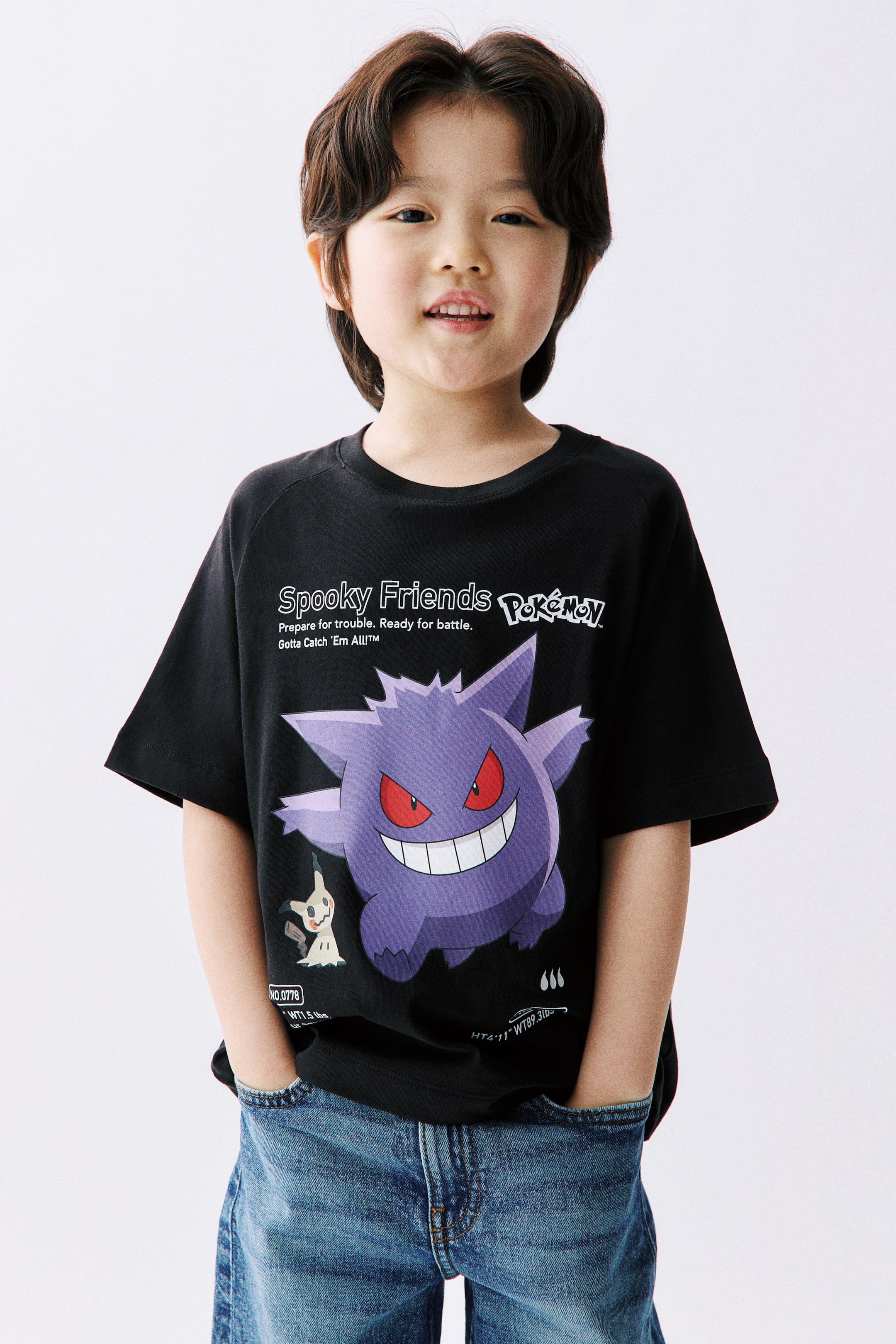 拡大画像を表示: 黒の半袖Tシャツには、大きな紫色のゲンガーキャラクターが赤い目と広い笑顔で描かれており、その横には小さな白いミミッキュキャラクターがいます。キャラクターの上には「Spooky Friends Pokémon」というテキストが印刷されており、キャラクターのステータスを詳細に示す小さなテキストが下にあります。