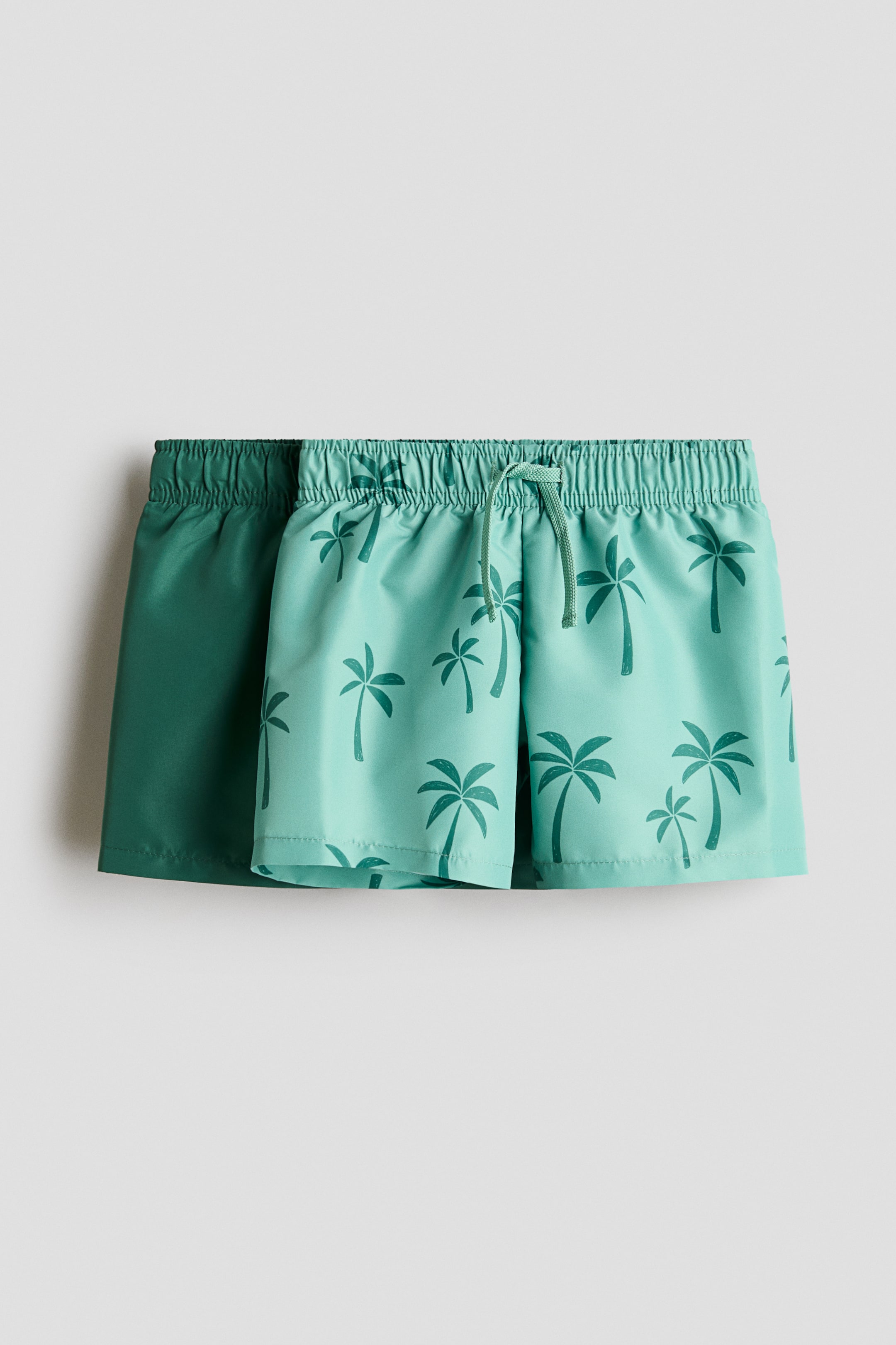 Agrandir l'image: Des shorts de bain sont présentés, avec deux paires montrées de face. La paire au premier plan est de couleur bleu sarcelle clair avec un motif de palmier et dispose d'une taille élastiquée avec un cordon de serrage. Une paire de couleur vert foncé uni est positionnée derrière, ayant également une taille élastiquée.