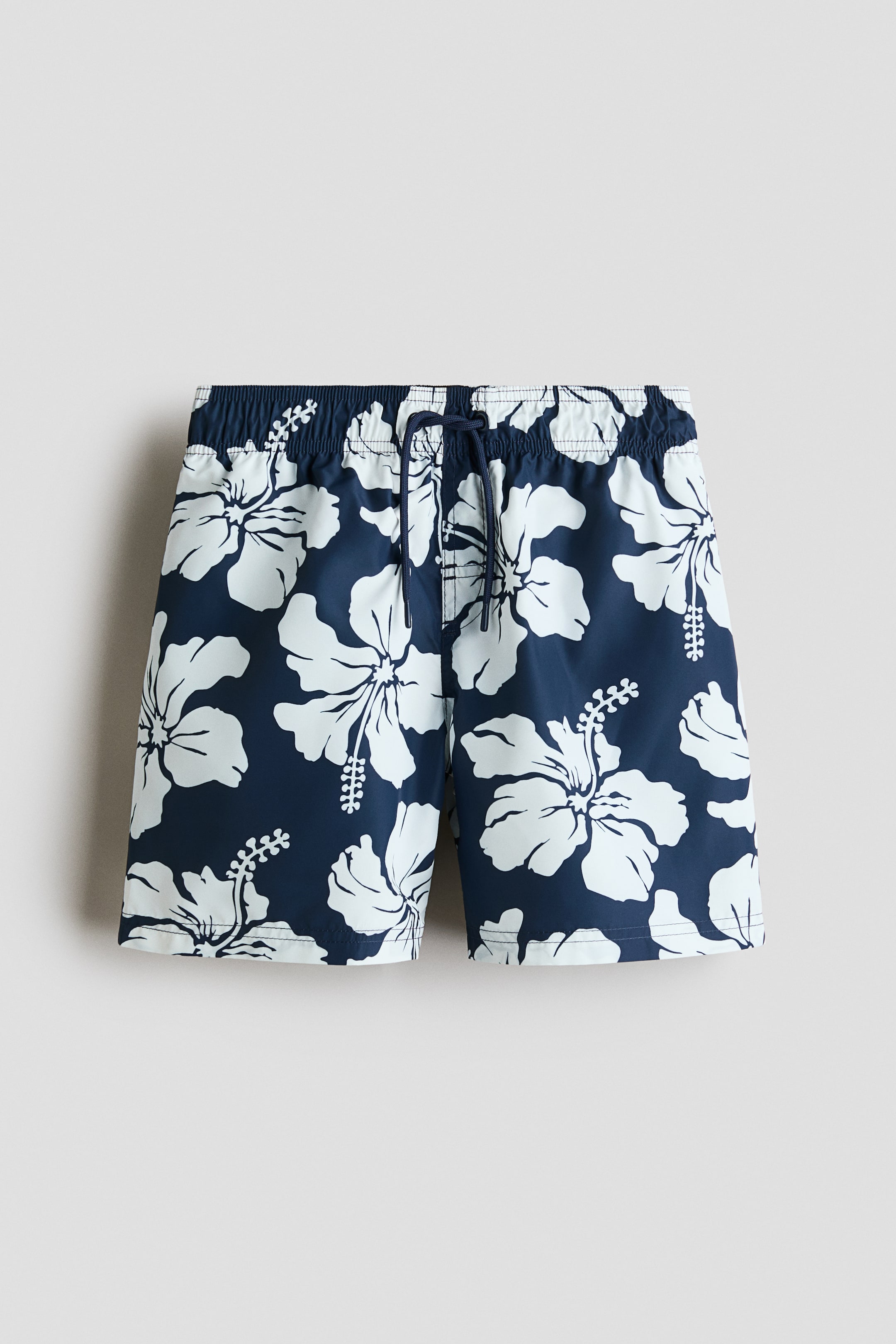 Größeres Bild ansehen: Bedruckte Badeshorts präsentieren sich in einem dunkelblauen Stoff mit großen weißen Hibiskusblumenmustern und einem elastischen Bund mit einer dunkelblauen Kordel, die vorne gebunden ist. Die Shorts sind mit einem Fake-Schlitz, einer aufgesetzten Gesäßtasche mit Haken- und Schlaufenverschluss sowie weichen Mesh-Innenshorts gestaltet.