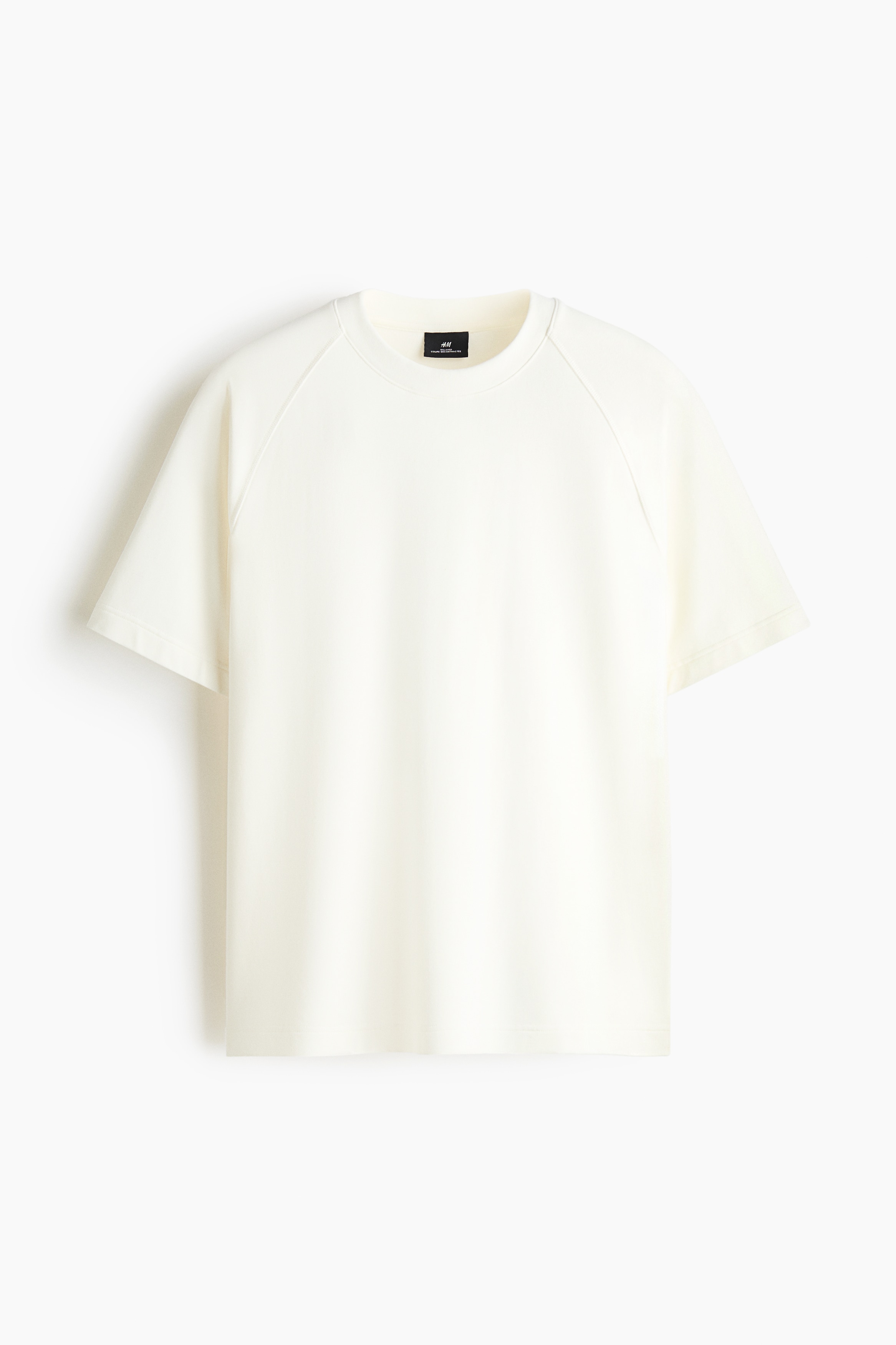 Un t-shirt en jersey Ã©pais de couleur crÃ¨me, avec col rond cÃ´telÃ© et manches raglan courtes, est portÃ© par le mannequin par-dessus une chemise blanche Ã  manches longues. Le t-shirt Ã  coupe dÃ©contractÃ©e prÃ©sente un ourlet droit, tandis que le mannequin repose ses mains prÃ¨s de ses hanches.
