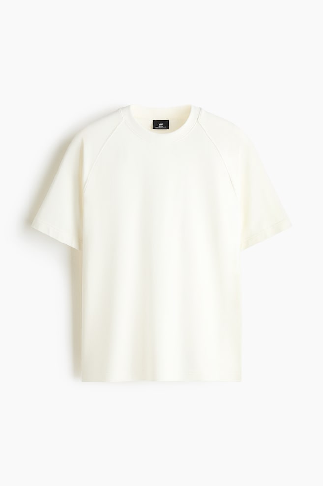 A cream-coloured, heavyweight jersey T-shirt is hanging front-facing, showcasing a relaxed fit, a round rib-trimmed neckline, short raglan sleeves, and a straight-cut hem. - 1