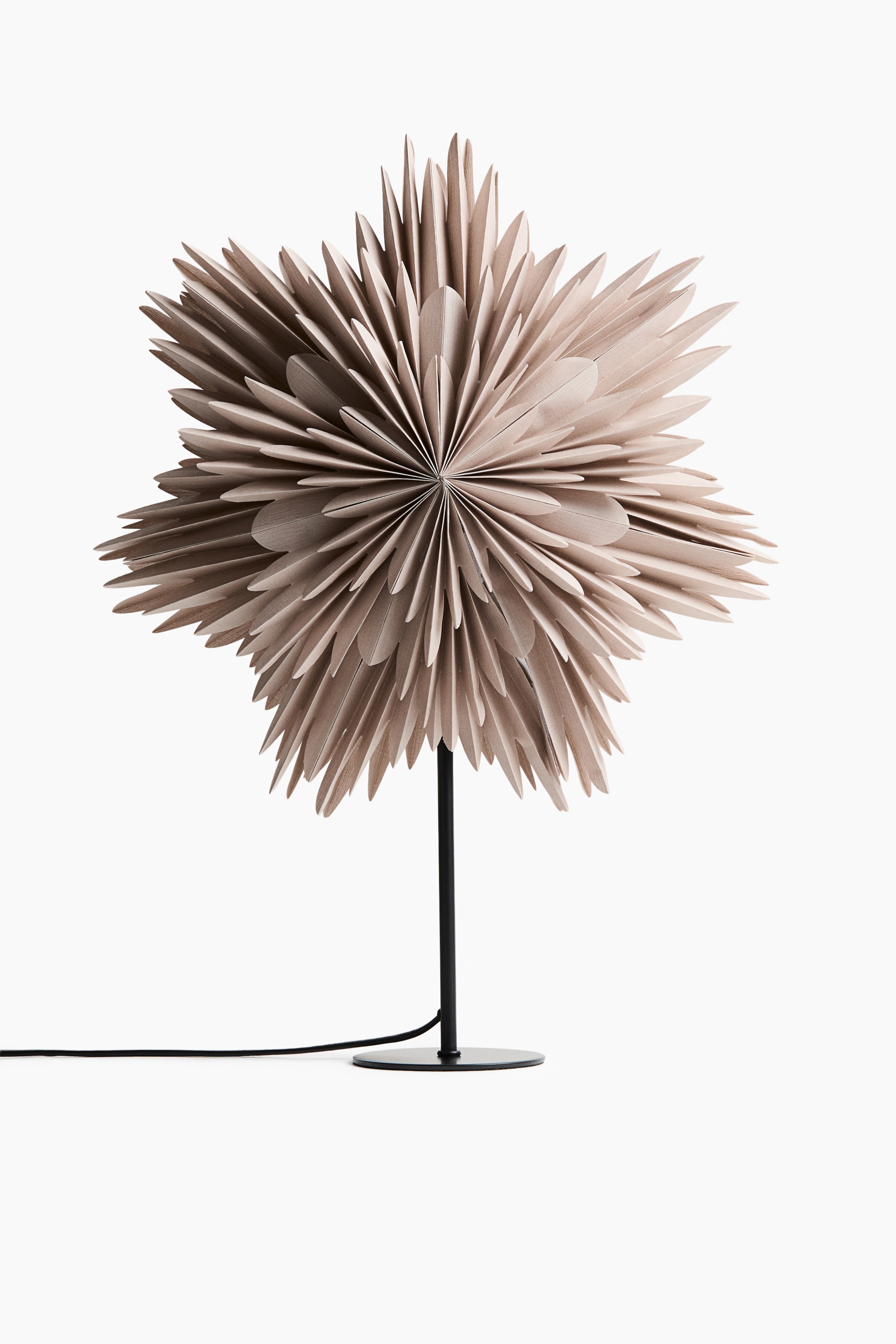 View larger image: A table lamp is shown facing forward, featuring a large, multi-layered beige paper shade shaped like a star, with numerous pointed folds radiating from the centre. The shade sits on a slender black metal stem rising from a flat circular base, with a black power cord extending to the side.