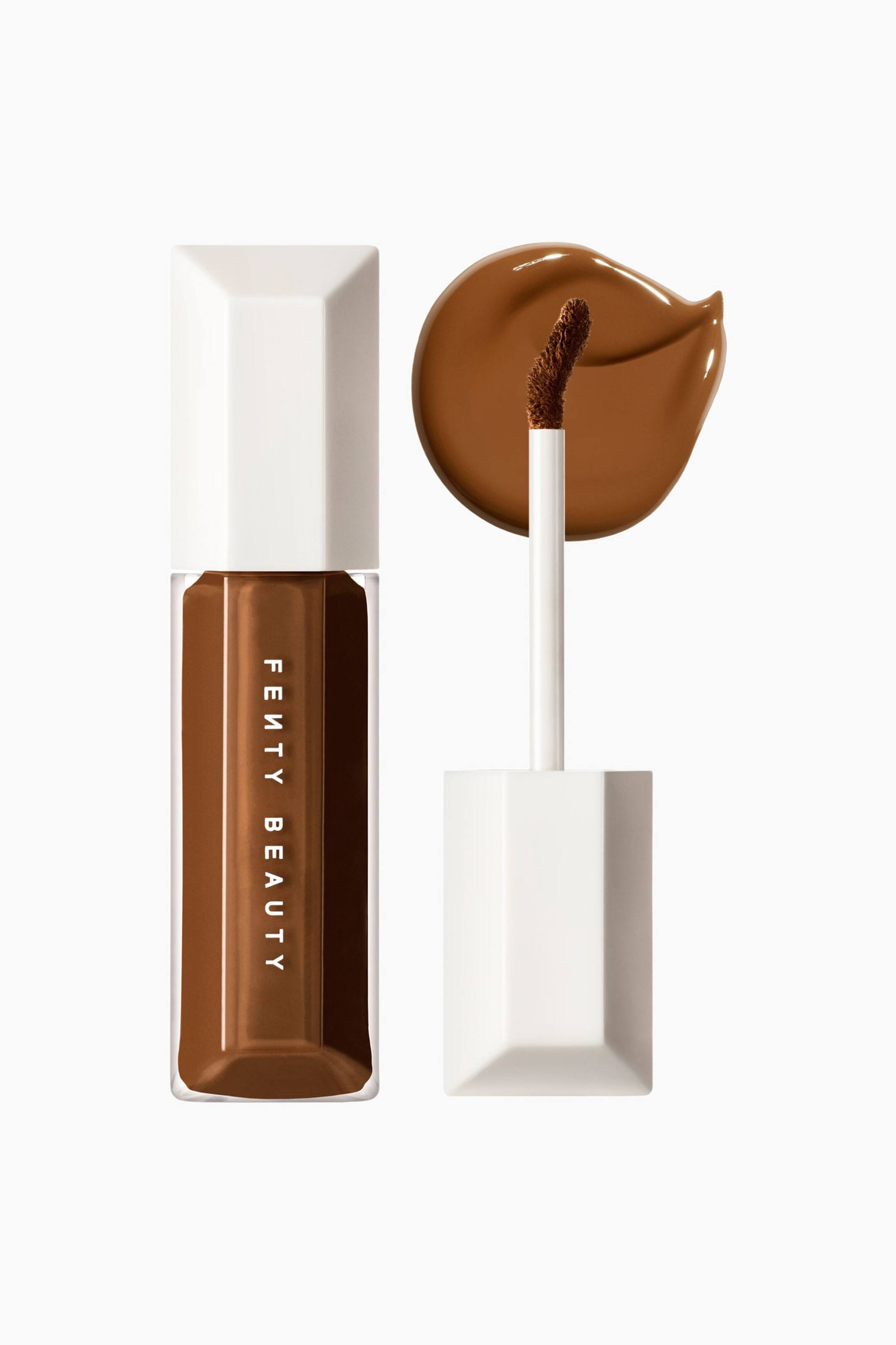 Visa större bild: We're Even Hydrating Longwear Concealer står upprätt i en klar sexkantig tub med vit kork, där den djupbruna formulan syns inuti. Bredvid den visas den avskruvade applikatorn upp med en böjd, vinklad spets, och bakom den syns ett slätt, glansigt runt swatch av concealern.