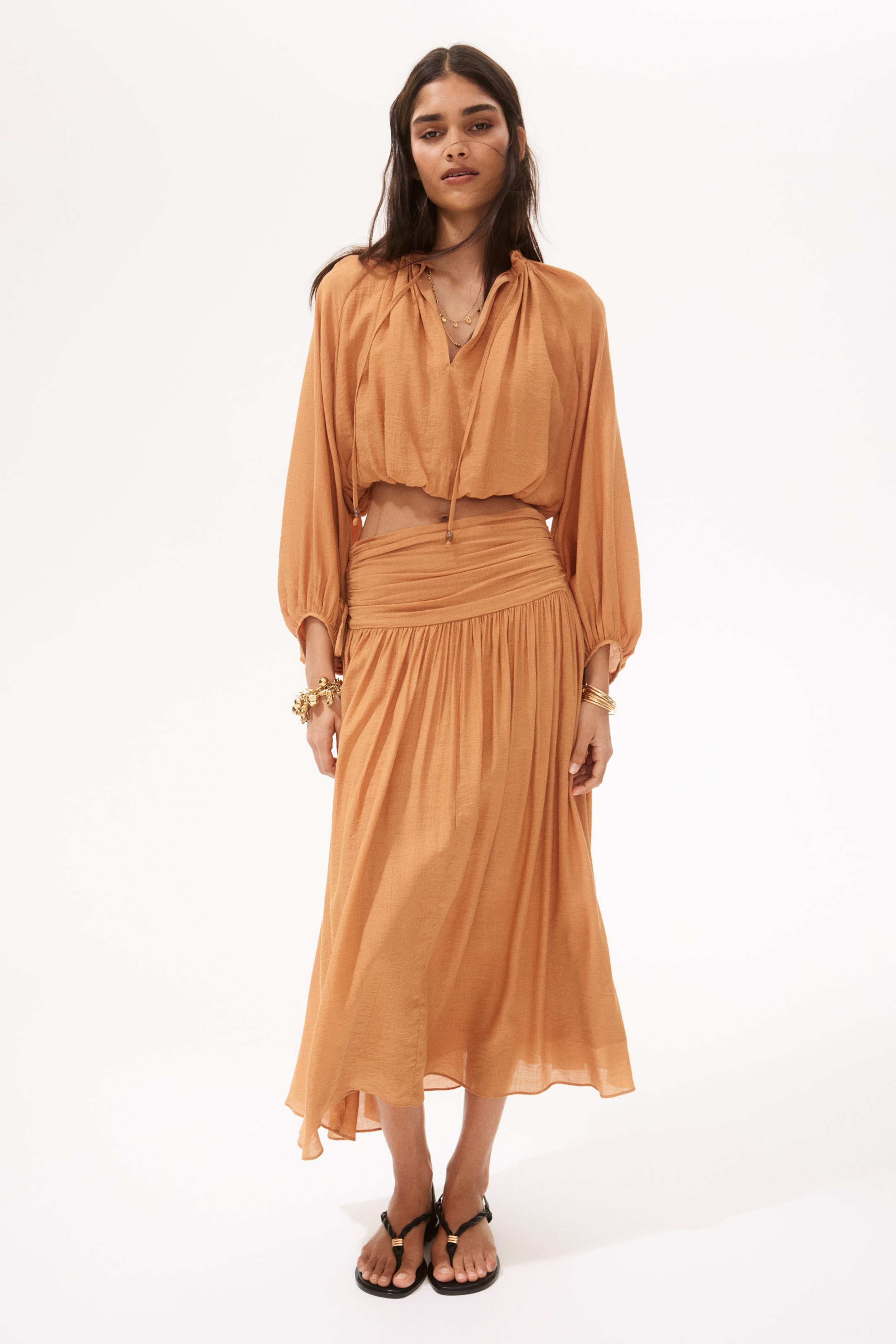 View larger image: The product is a subtle orange two-piece ensemble, comprising a cropped top and a midi skirt, displayed from a front-facing perspective. The top highlights a v-neckline with ties, long balloon sleeves with elasticated cuffs, and gathered detailing, while the skirt showcases a ruched waistband and a flowing silhouette in a textured fabric.