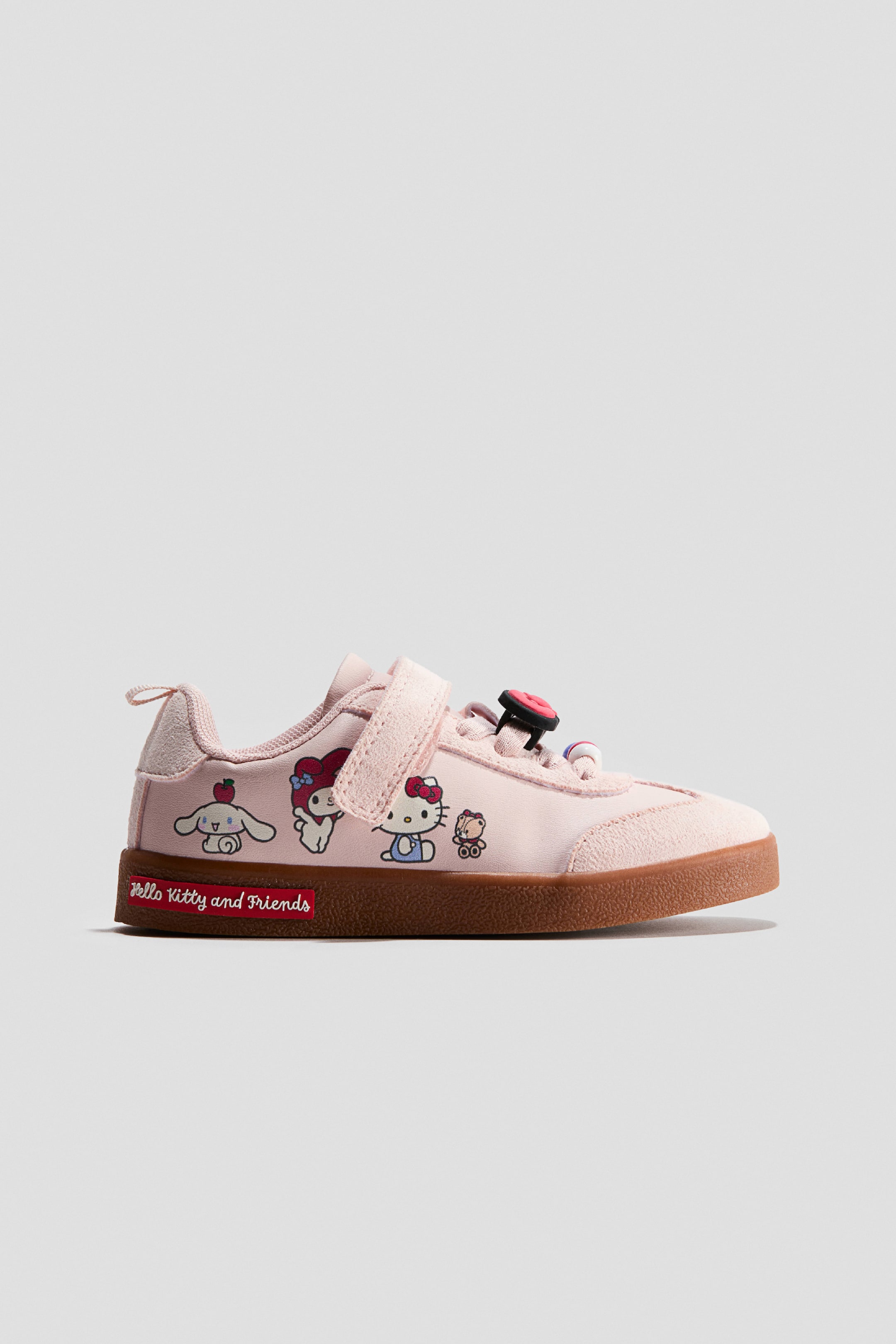 View larger image: A light pink children's sneaker, shown in side profile, features printed illustrations of Hello Kitty and Friends characters along the side. The shoe has a hook-and-loop strap closure, textured suede overlays on the toe and heel, and a brown gum sole with a red logo label.