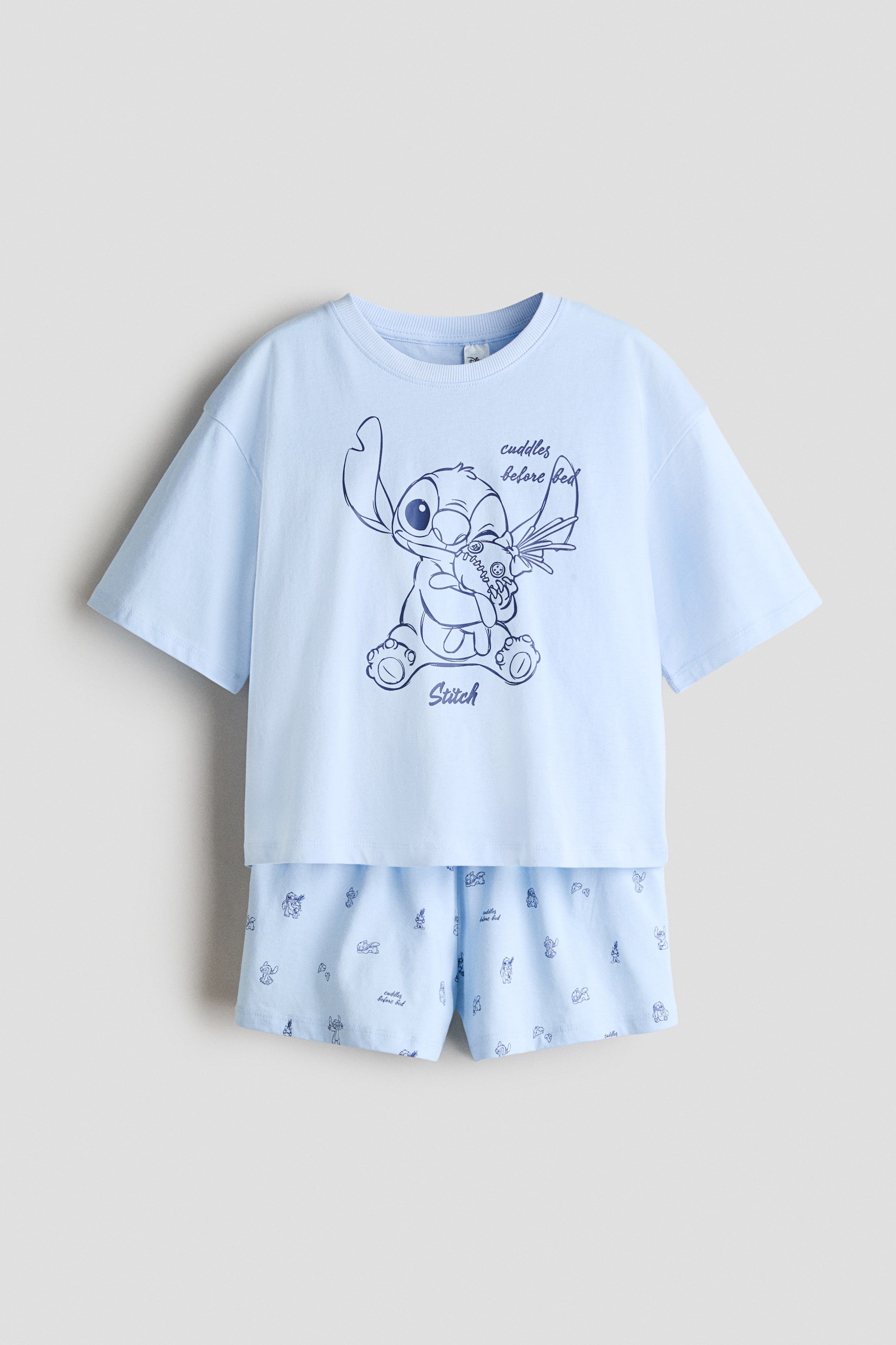 View larger image: A light blue two-piece pajama set features a short-sleeved top with a ribbed crew neck, adorned with a large dark blue graphic of Stitch holding a plush toy, along with the phrases "cuddles before bed" and "Stitch". The matching shorts showcase an all-over small print of Stitch in various poses, interspersed with the text "cuddles before bed".