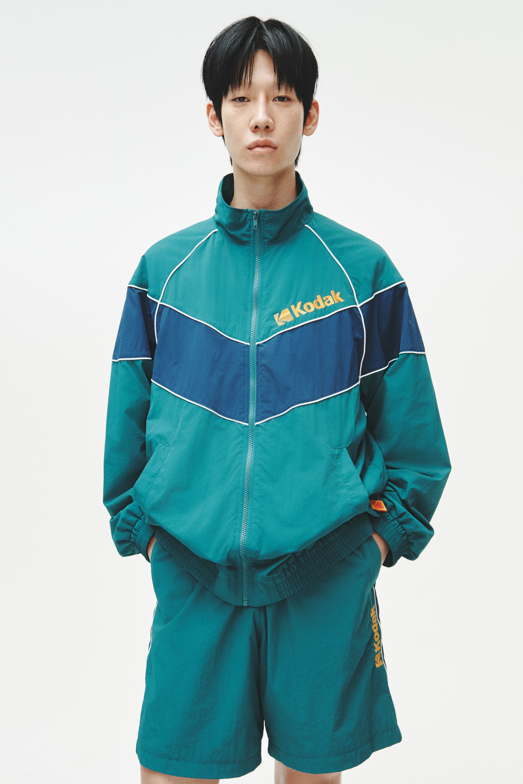 View larger image: A teal green at navy blue na tracksuit ay nagtatampok ng isang harapang full-zip jacket na may stand-up collar, puting piping na nagbabalangkas ng navy blue na chevron stripe, isang orange na Kodak logo sa kanang bahagi ng dibdib, at elasticated cuffs at hem. Ang katugmang teal green na shorts ay nagtatampok ng orange na Kodak logo at isang puting side stripe sa kaliwang binti.