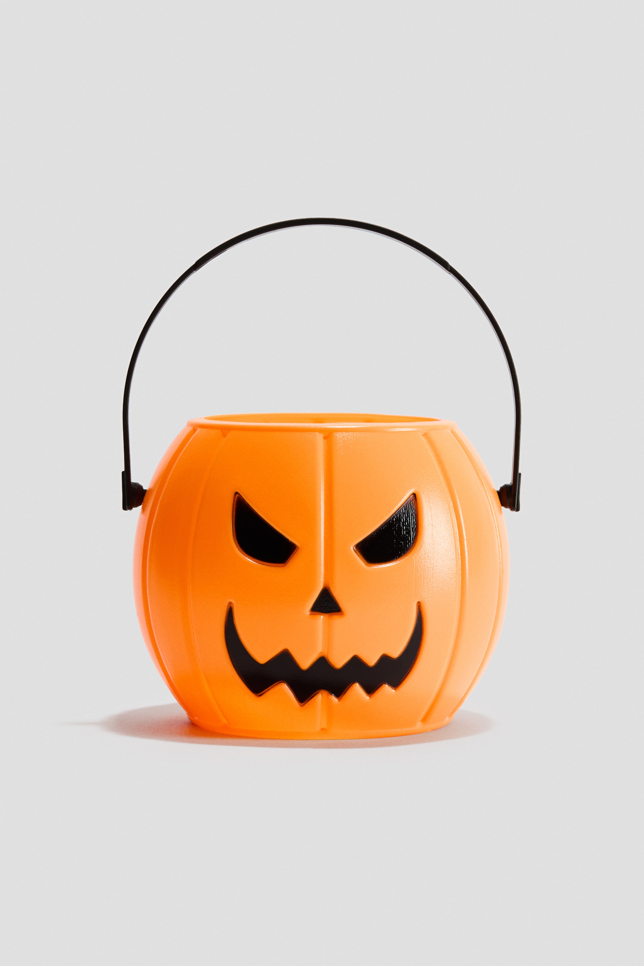 View larger image: An orange plastic pumpkin-shaped bucket features a carved black face design, including angular eyes and a jagged smile. A black plastic handle arches over the top of the bucket.