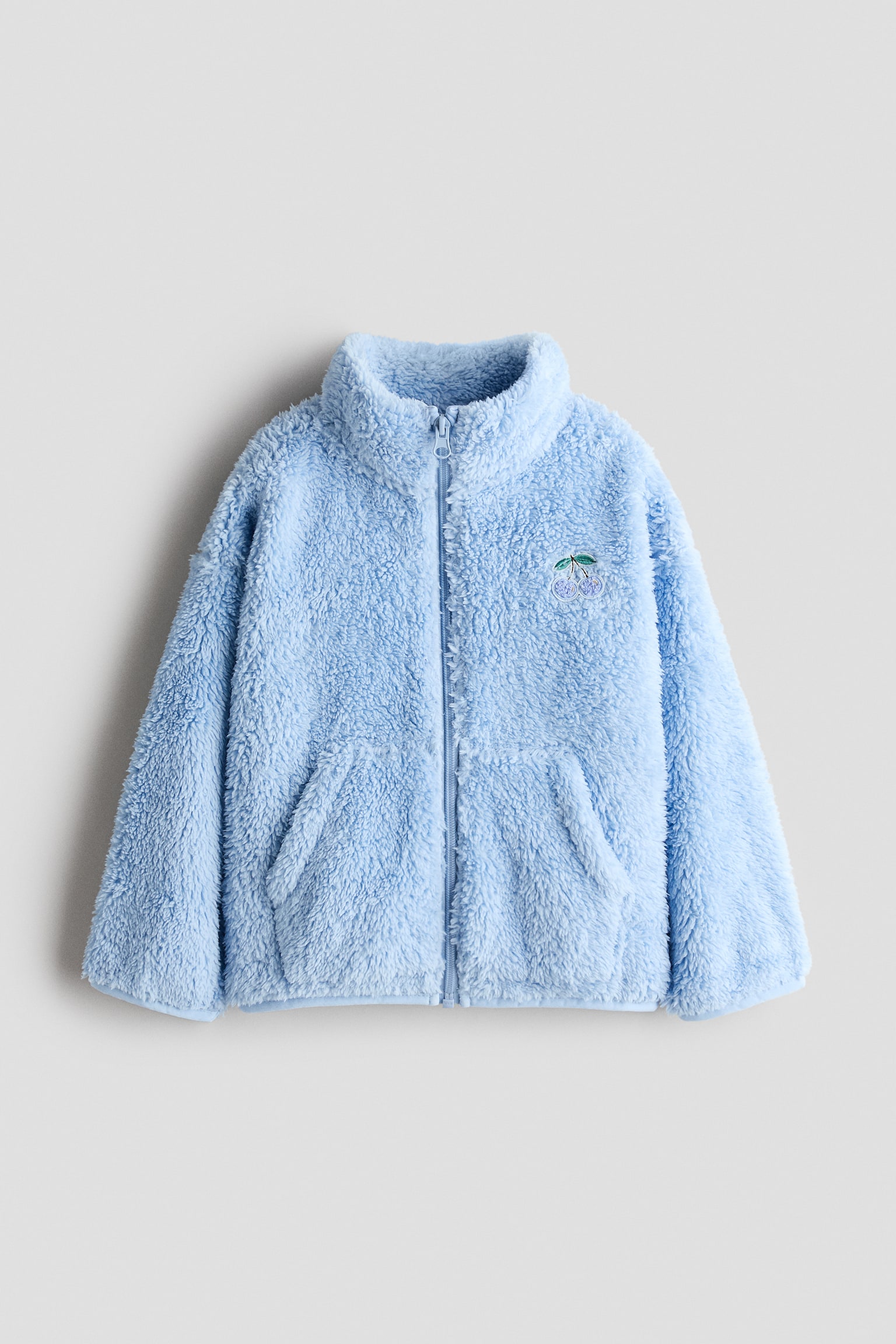 A light blue, fluffy fleece jacket is shown from the front, featuring a full-length zip fastening, a high stand-up collar, and two front slip pockets. A small embroidered cherry detail in lilac and green decorates the left chest.