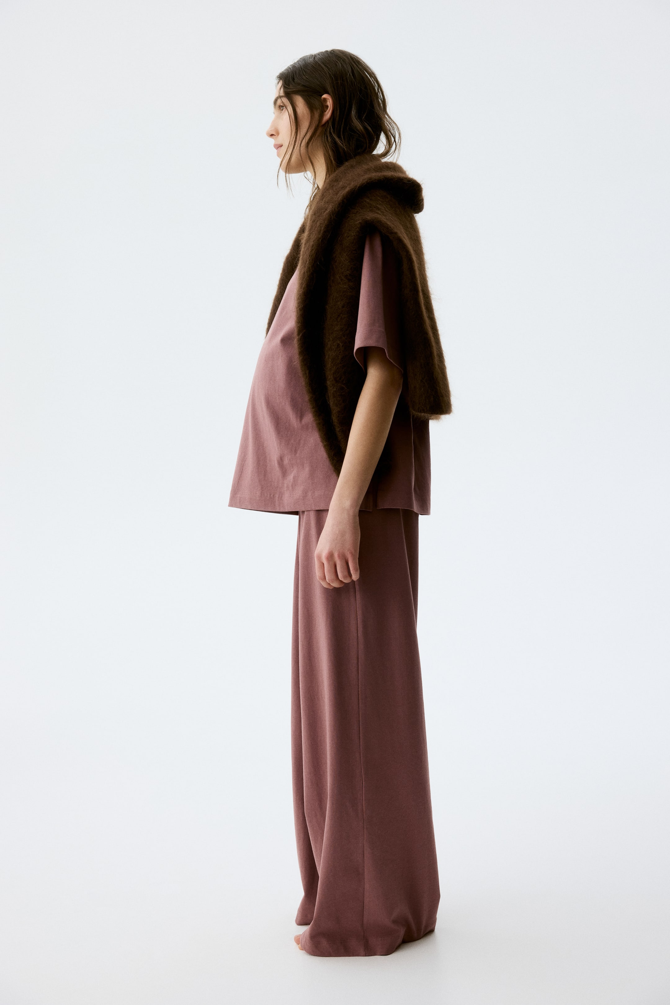 View larger image: A two-piece ensemble, showcasing a loose-fitting, short-sleeved top paired with matching wide-leg, full-length trousers in a dusty rose hue, is displayed from a side profile. A dark brown, textured shawl is elegantly draped over the shoulders and upper arms, layering over the top.