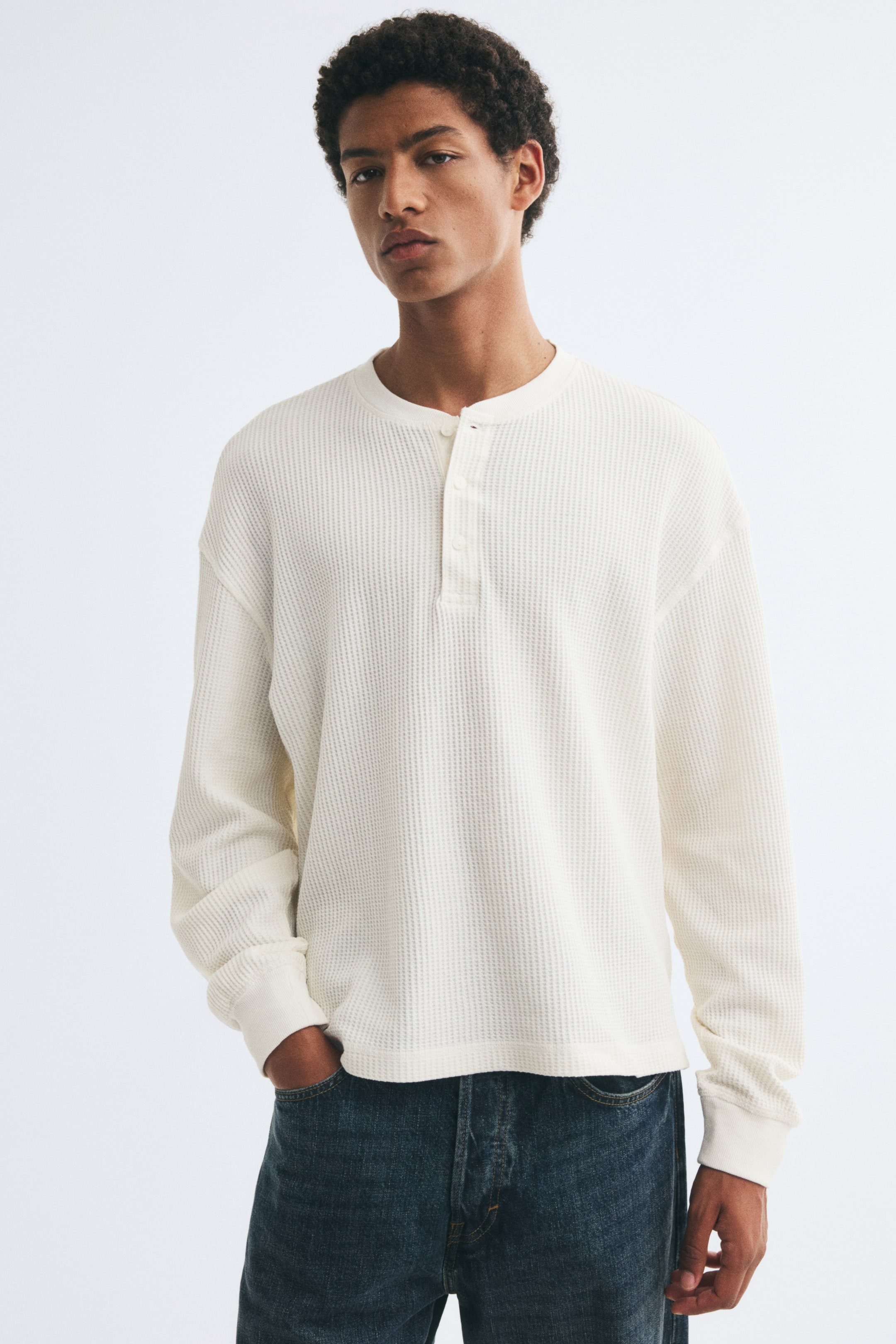 View larger image: The model is wearing a long-sleeve off-white waffle-knit jersey top featuring a round neckline with a button placket, dropped shoulders, and ribbed cuffs. The top is styled with dark blue denim jeans, and the model has one hand resting in a front pocket.