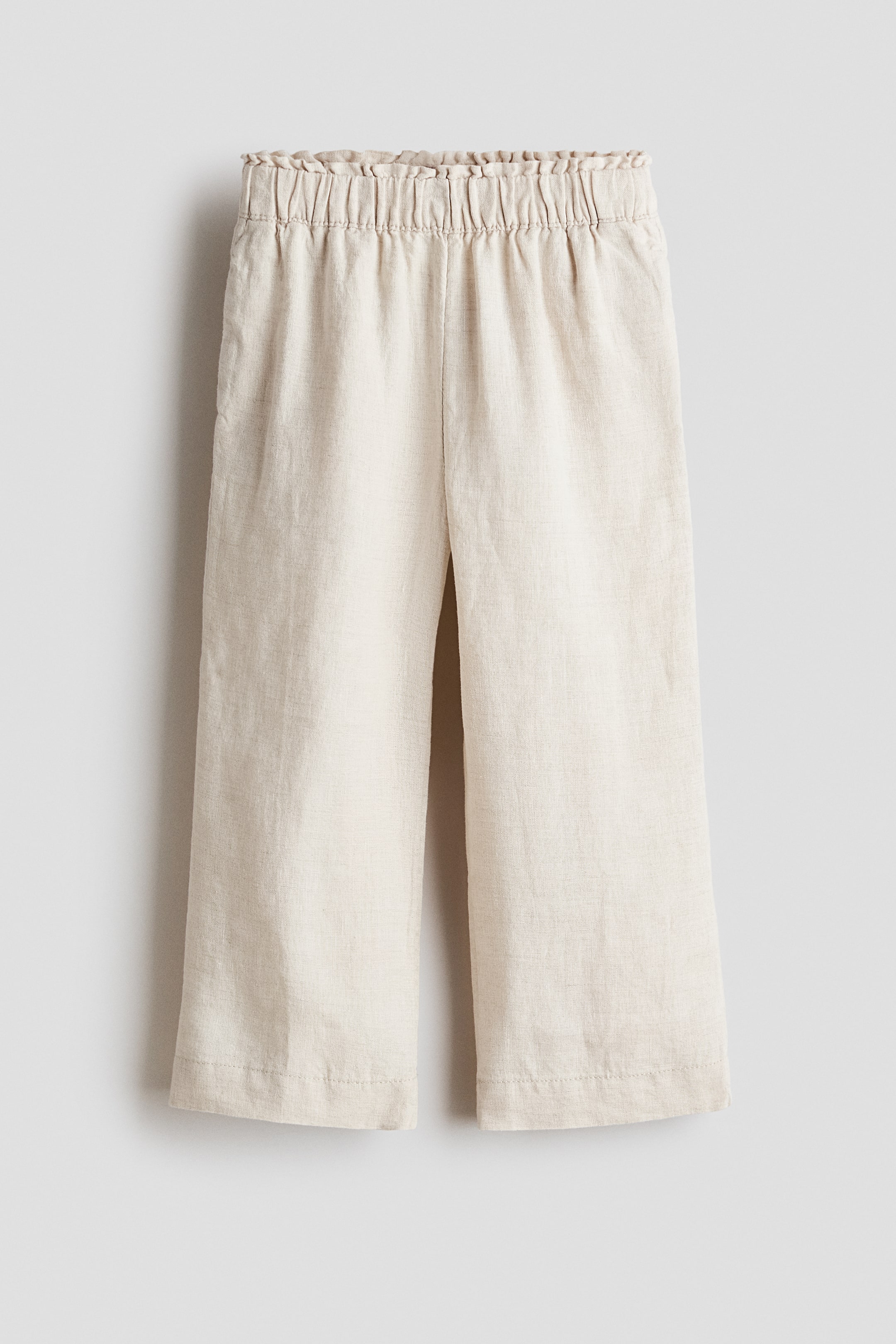 View larger image: Light beige, linen-textured pants shown from the front, featuring a ruffled elastic waistband and wide, cropped legs.