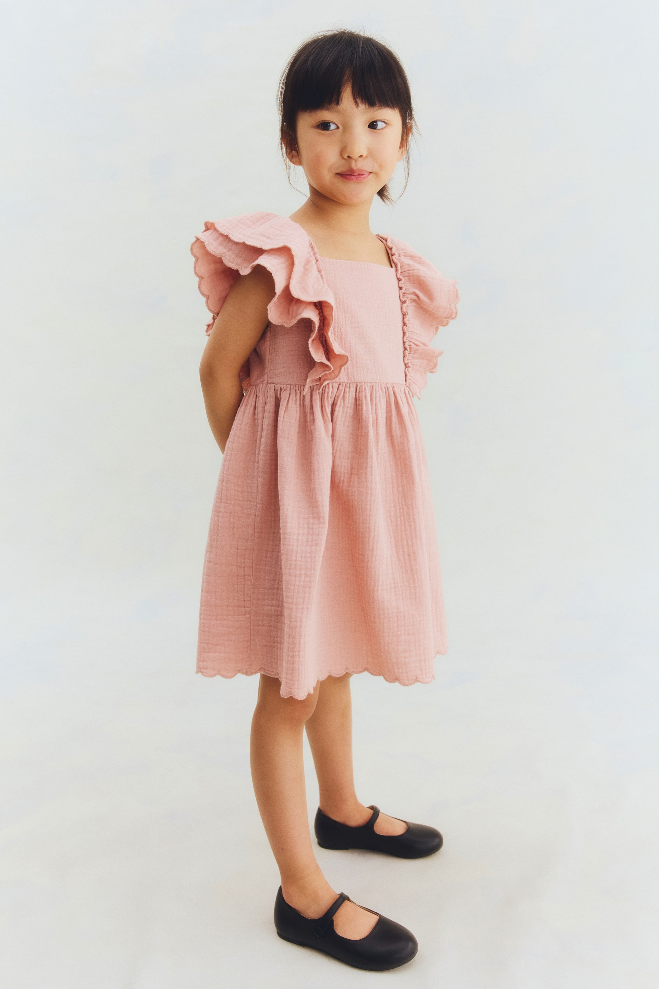 View larger image: A pink sleeveless dress crafted in a soft weave showcases its front, featuring a square neckline and a narrow-cut yoke. Double-layered frills with scalloped edges extend over the shoulders, while gathers appear at the waist above a flared skirt with a scalloped hem.