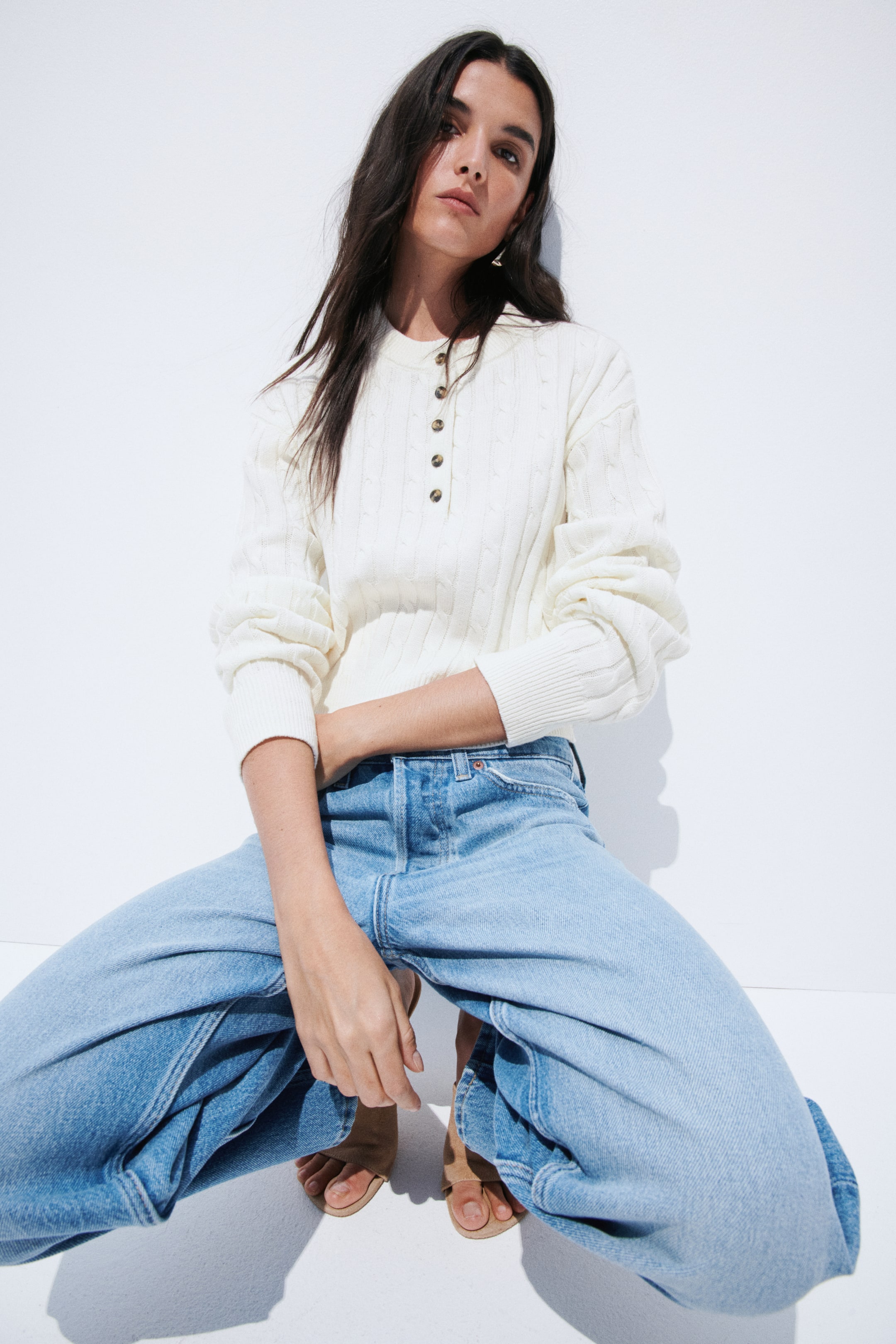 View larger image: A cream cable knit jumper is showcased with a relaxed fit, featuring a Henley placket with four brown buttons, a round neckline, and ribbed cuffs and hem.