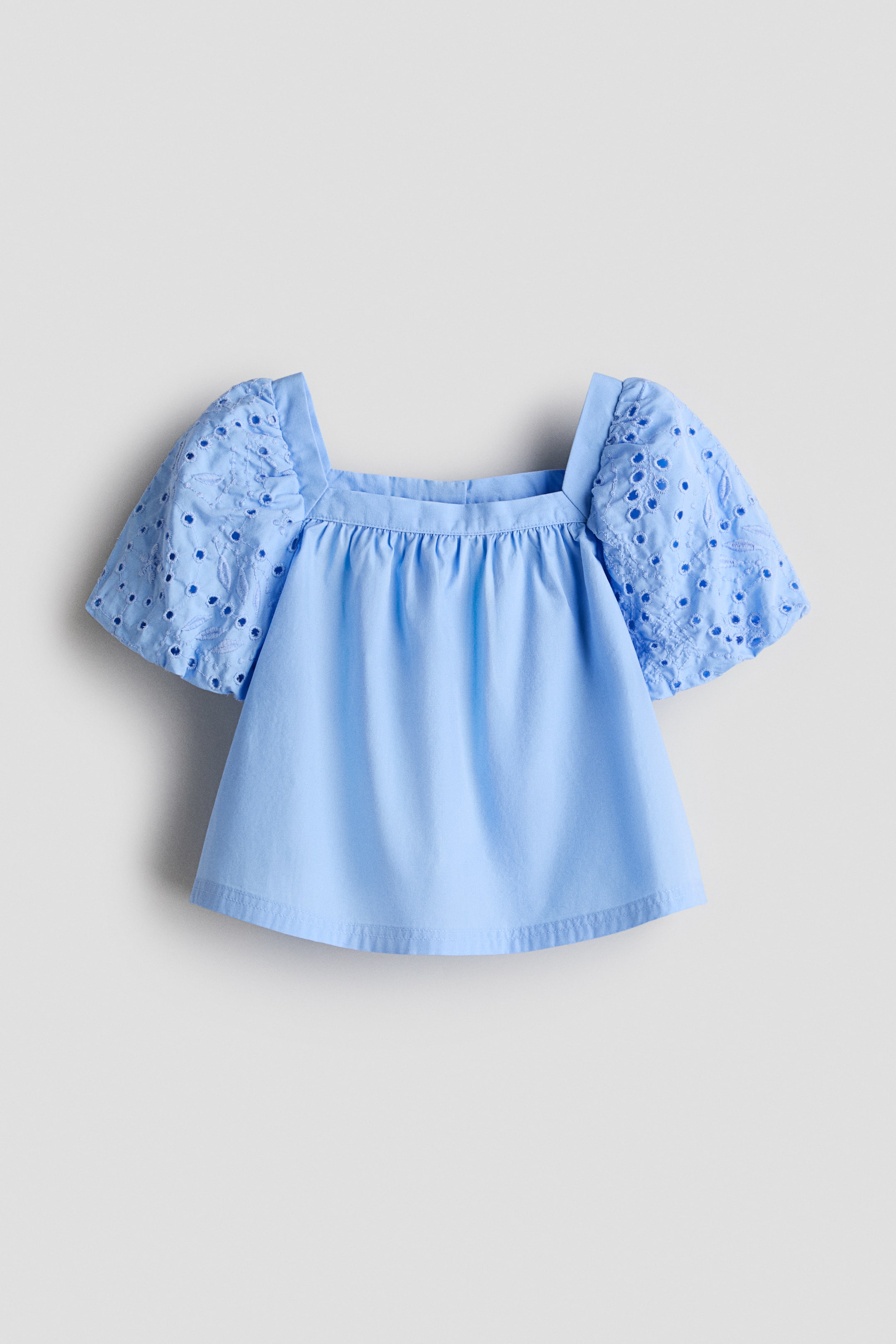 View larger image: A light blue top, shown from the front, is featuring a square neckline with gathering detail below it. Its short, puffed sleeves are displaying broderie anglaise embroidery.