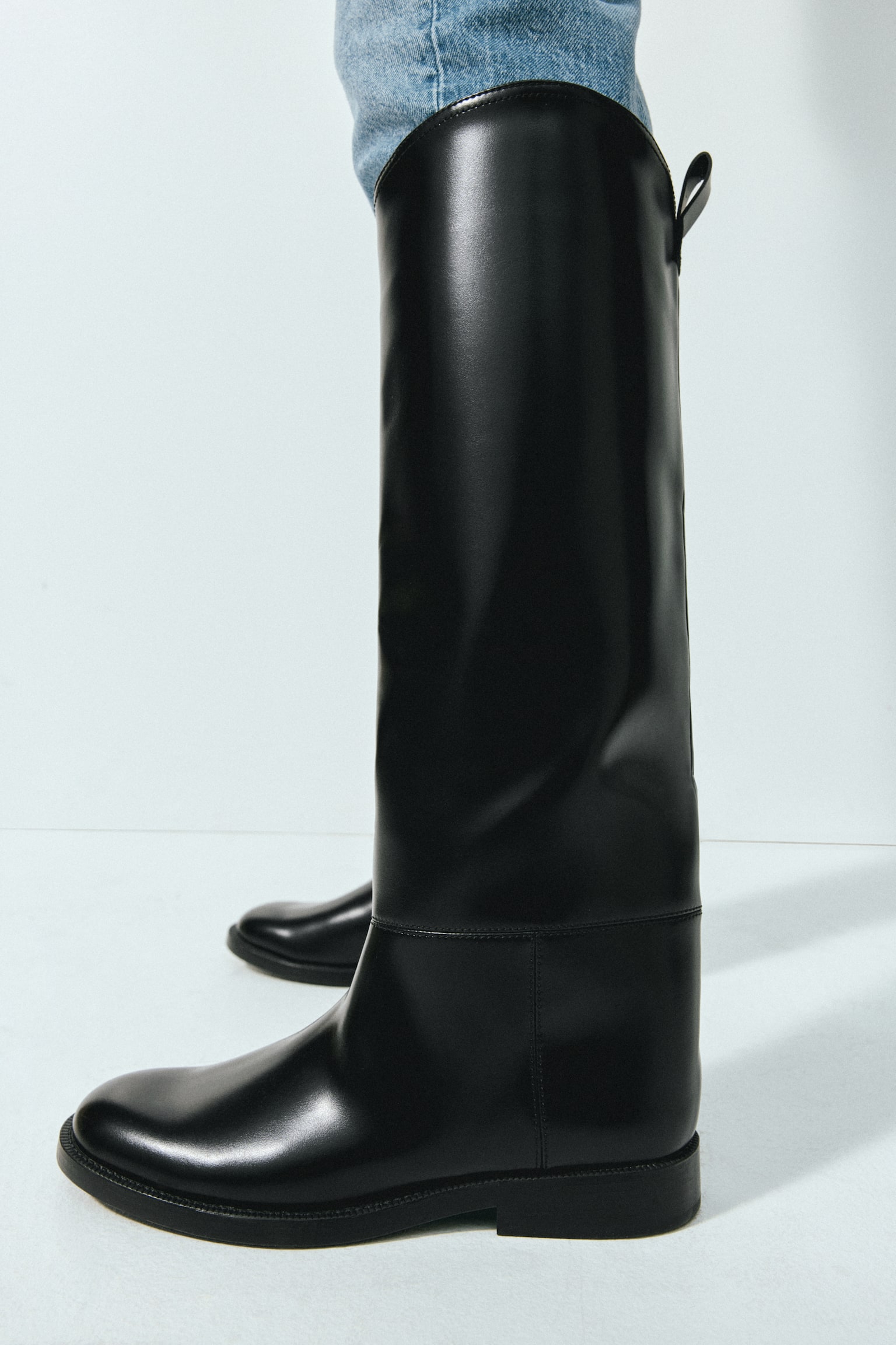 A black boot in coated fabric is displayed from a side angle, showcasing rounded toes, knee-high shafts, and a small loop at the back of the shaft. - 1