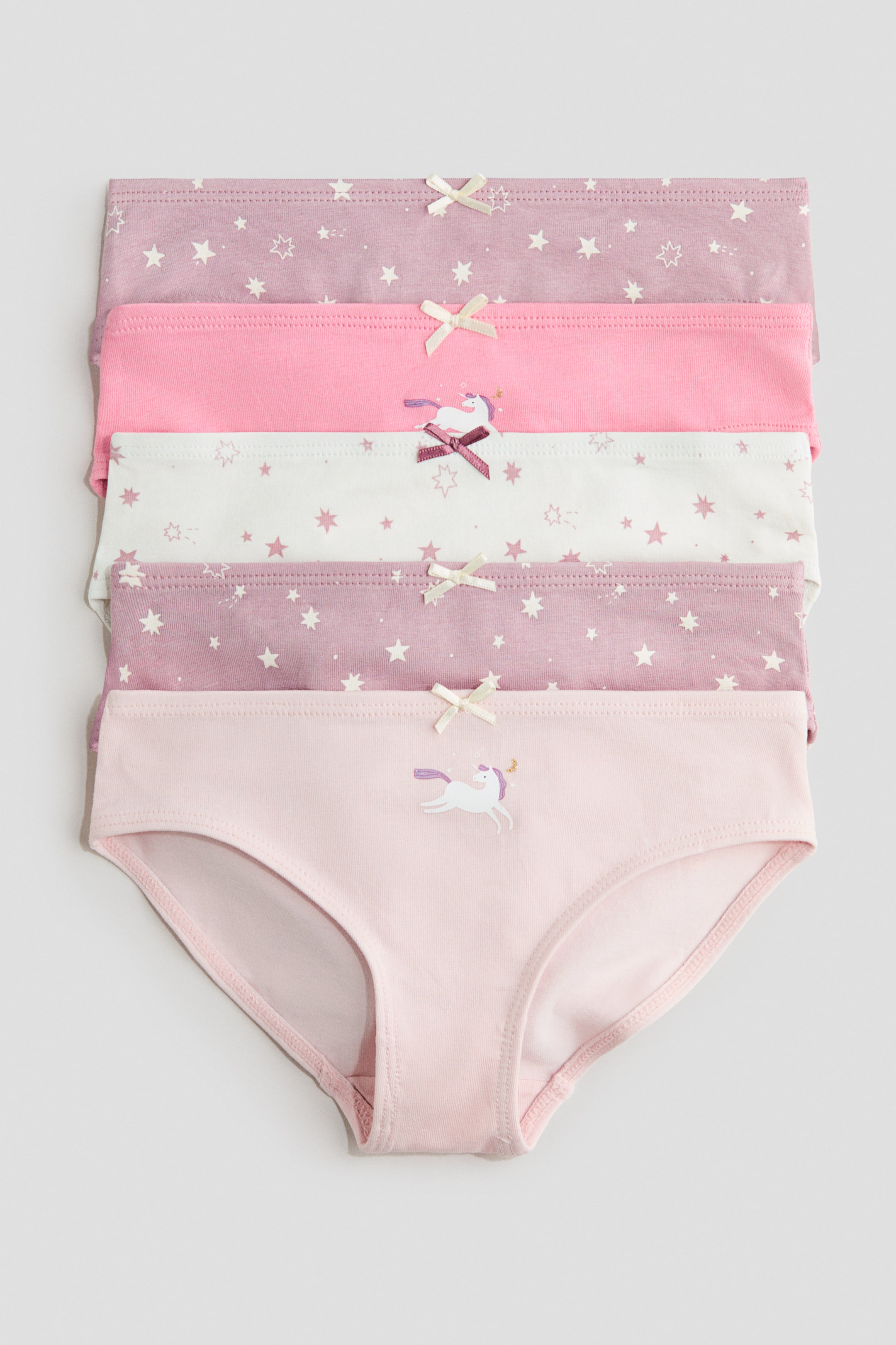 View larger image: Five pairs of children's underwear are displayed stacked horizontally, facing forward. The set includes two purple pairs with white star patterns and cream bows, one solid pink pair with a cream bow, and one white pair with purple stars and a unicorn graphic featuring a dark pink bow. The bottom, fully visible light pink pair features a unicorn graphic on the front and showcases its brief style.