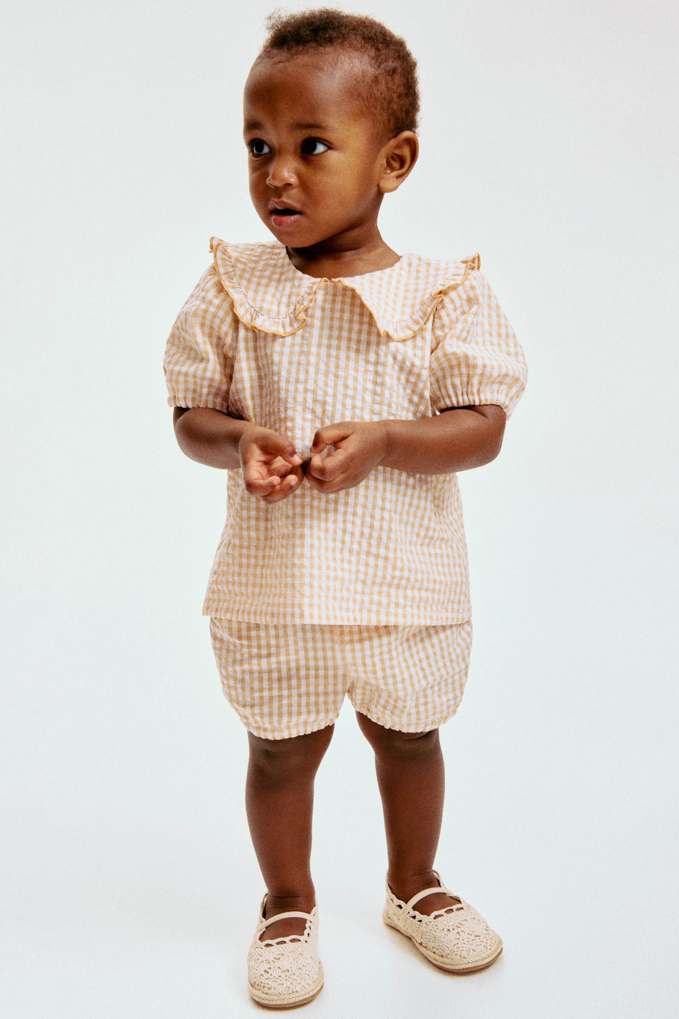 View larger image: Presenting a light brown and white gingham check two-piece set, featuring a top adorned with a ruffled Peter Pan collar and puffed sleeves, paired with bloomer shorts that have a gathered waist and leg openings. The ensemble is styled with cream crocheted Mary Jane shoes, complete with an espadrille-style sole and a scalloped strap.