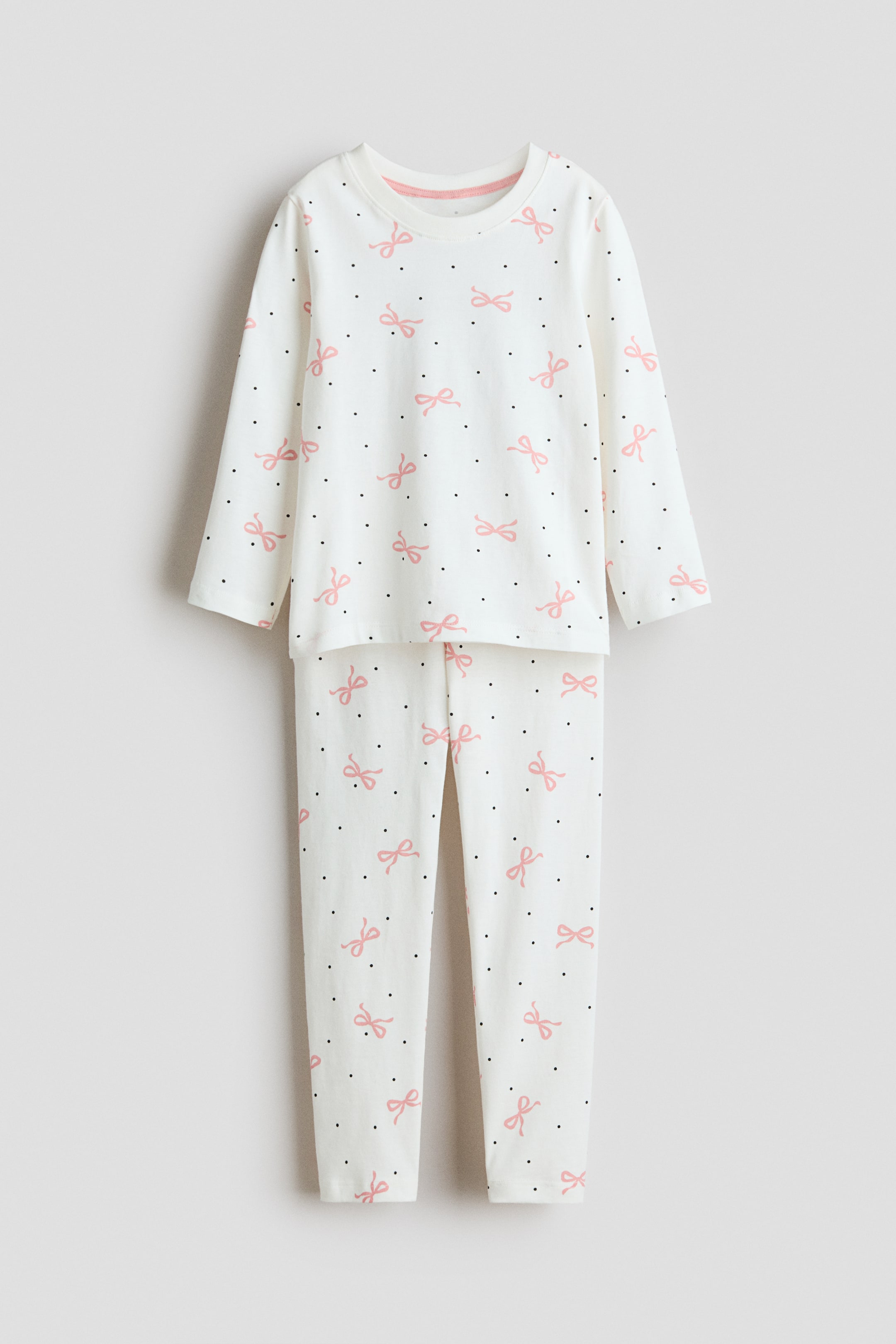 View larger image: A pair of pyjamas is being presented FRONT, consisting of a long-sleeved top with a round neckline and matching full-length bottoms. Both pieces are featuring a white base fabric printed with a pattern of small pink bows and tiny black dots.