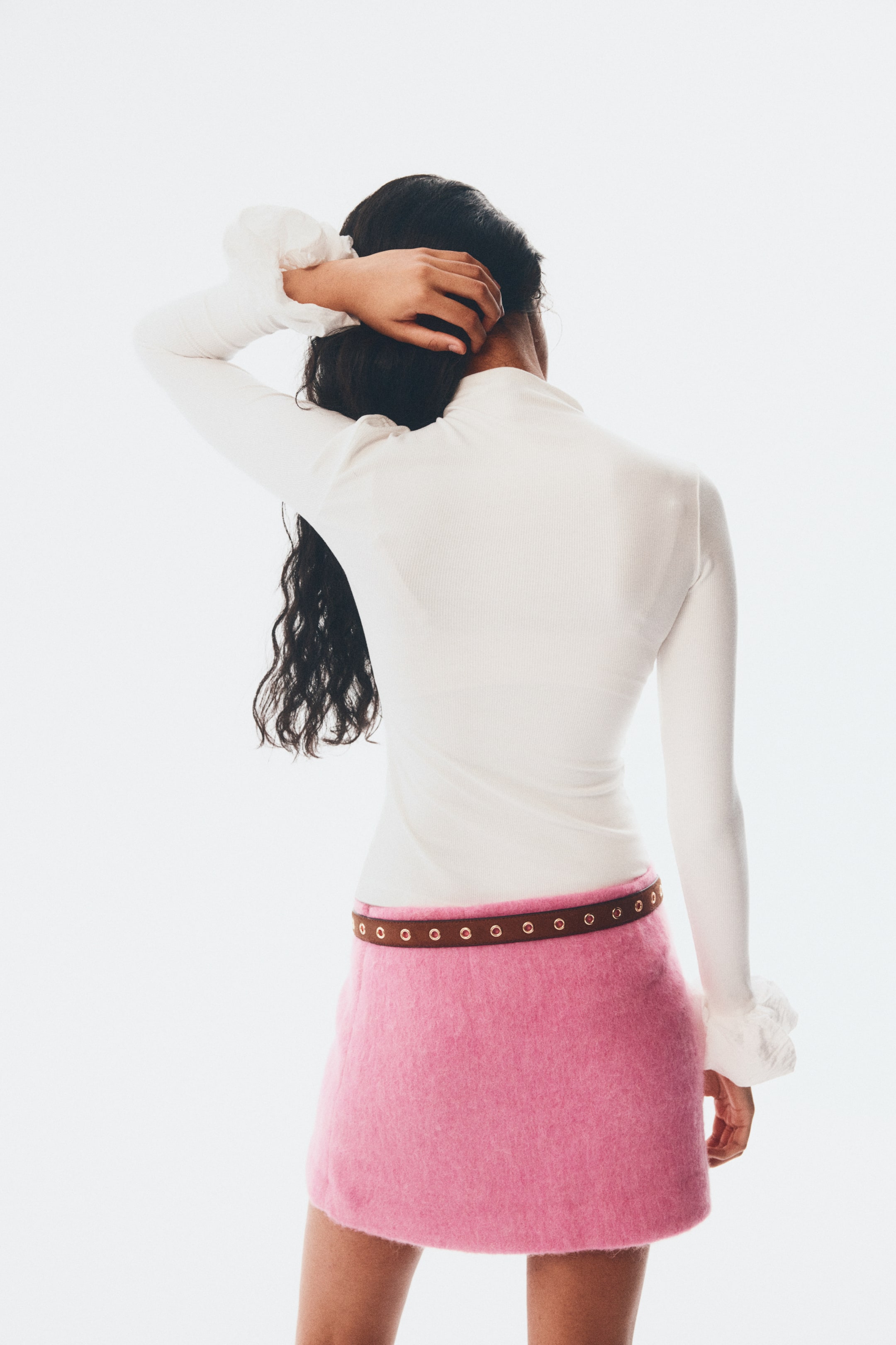 View larger image: A white ribbed long-sleeve top, featuring ruffled cuffs and a mock neck, is visible from the back. It is styled with a pink textured mini skirt and a brown belt featuring metal eyelets.