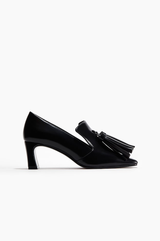 Ein schwarzer Lackleder-Loafer-Pump präsentiert eine spitze Zehenpartie, einen niedrigen ausgestellten Blockabsatz und ein großes Quastendetail, das den Rist schmückt. - 1