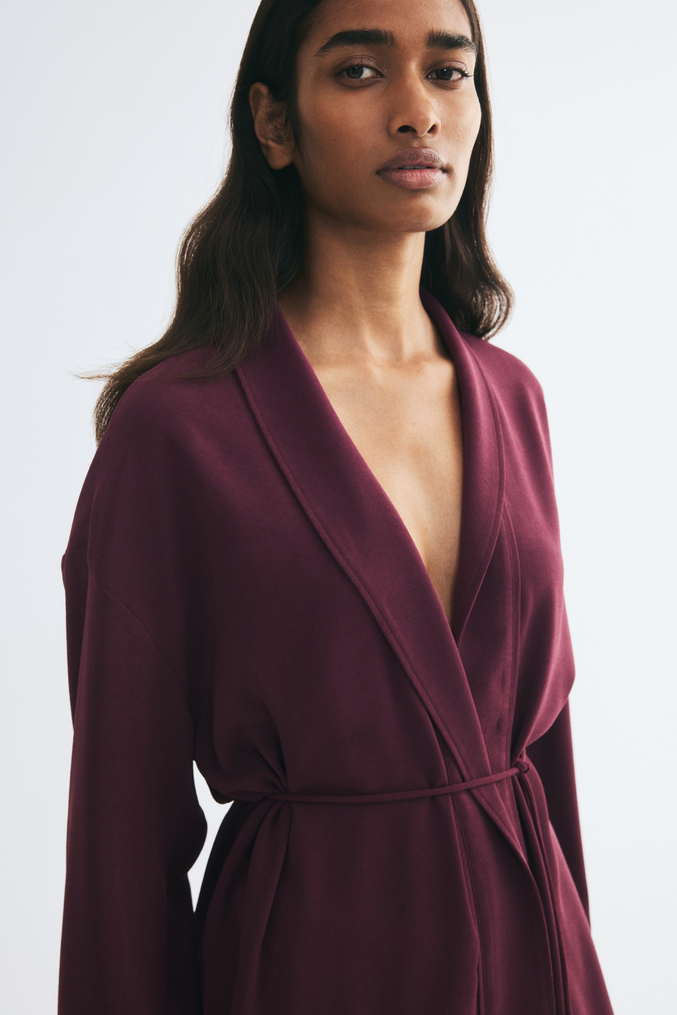 A deep plum wrap jacket is hanging front-facing, showcasing an open-front design with a draped shawl collar. The jacket features dropped shoulders, wide-cuffed sleeves, and a thin, tasseled drawstring tie at the waist.