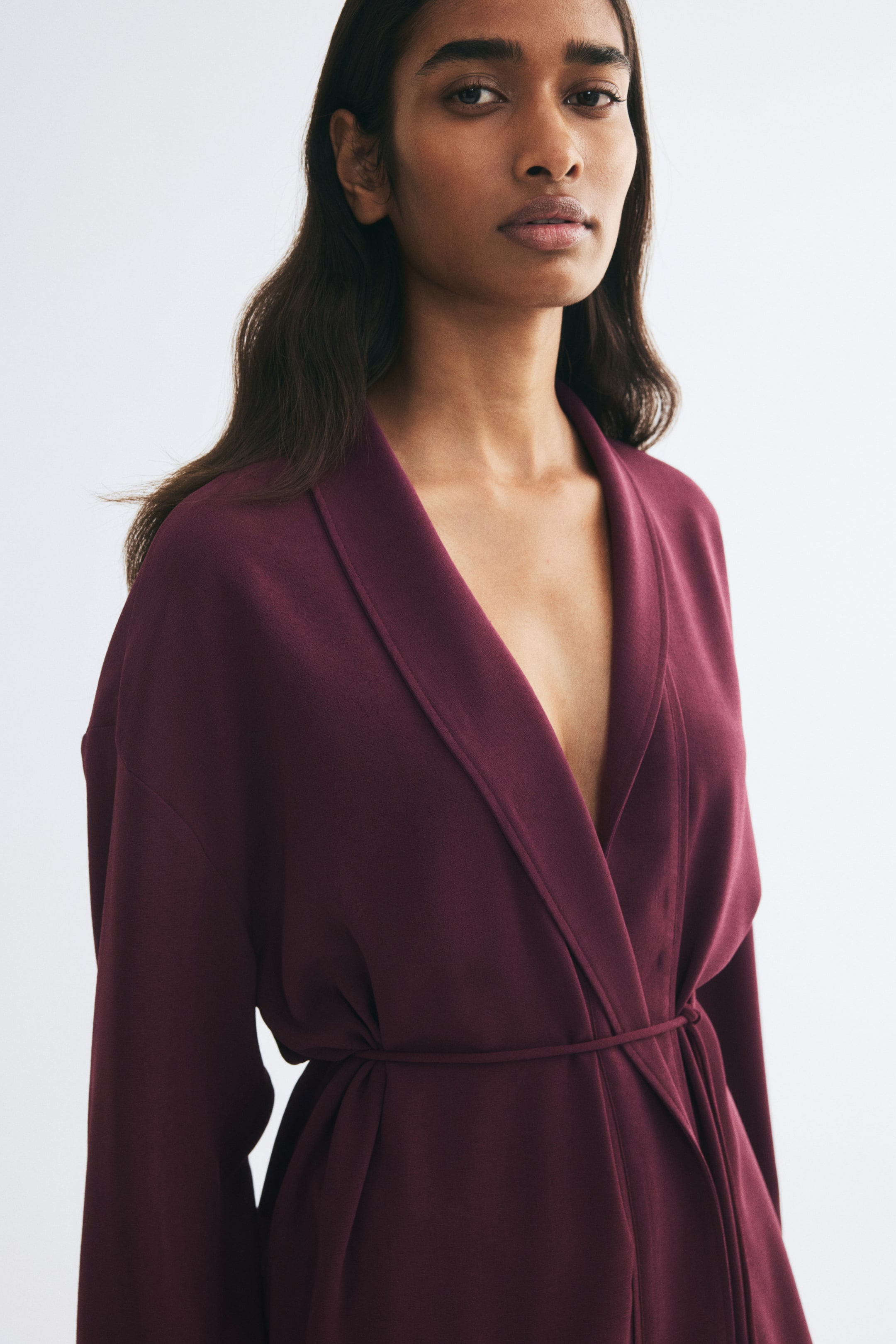 View larger image: A deep burgundy wrap coat featuring a wide shawl collar and a thin self-tie belt is worn by the model. The garment has dropped shoulders and creates a deep V-neckline.