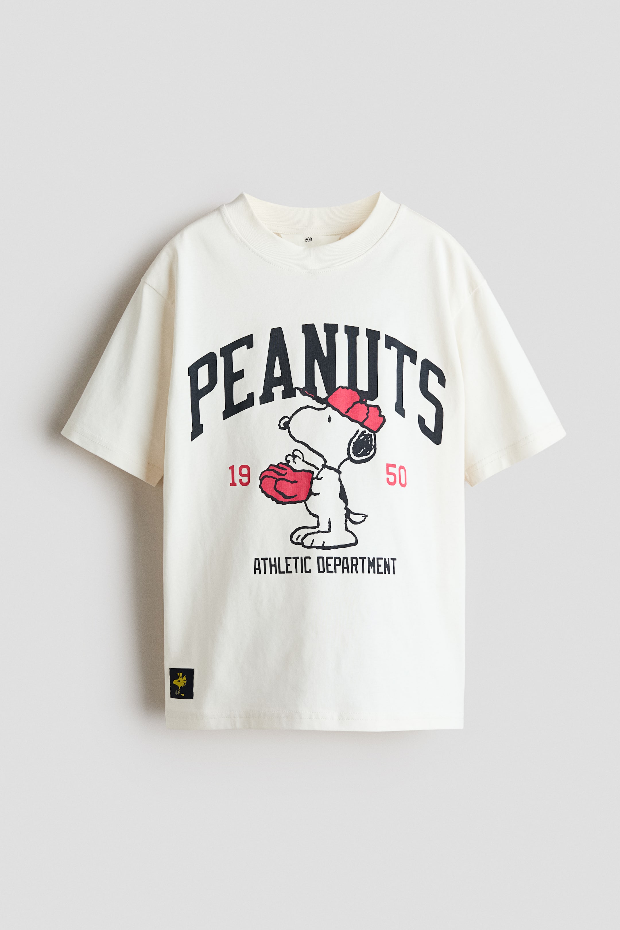 View larger image: A cream T-shirt showcases its front, featuring a large graphic with "PEANUTS" in dark arched lettering above Snoopy, who is wearing a red cap and glove. The design is flanked by red "19" and "50," with "ATHLETIC DEPARTMENT" below. The T-shirt is crafted with a ribbed crew neck, dropped shoulders, and a loose fit.