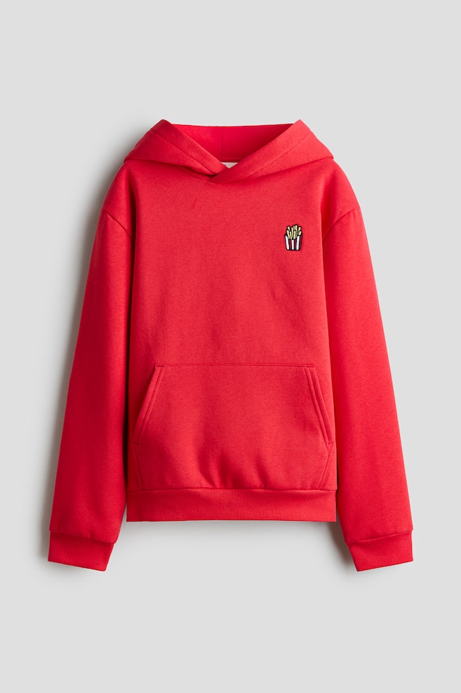 Czerwona bluza z kapturem typu pullover jest pokazana z przodu, z maÅÄ
haftowanÄ
naszywkÄ
przedstawiajÄ
cÄ
frytki w paskowanym pojemniku na lewej piersi. Ma klasycznÄ
kieszeÅ kangurkowÄ
, dÅugie rÄkawy ze ÅciÄ
gaczowymi mankietami oraz ÅciÄ
gaczowy dóÅ. - 1