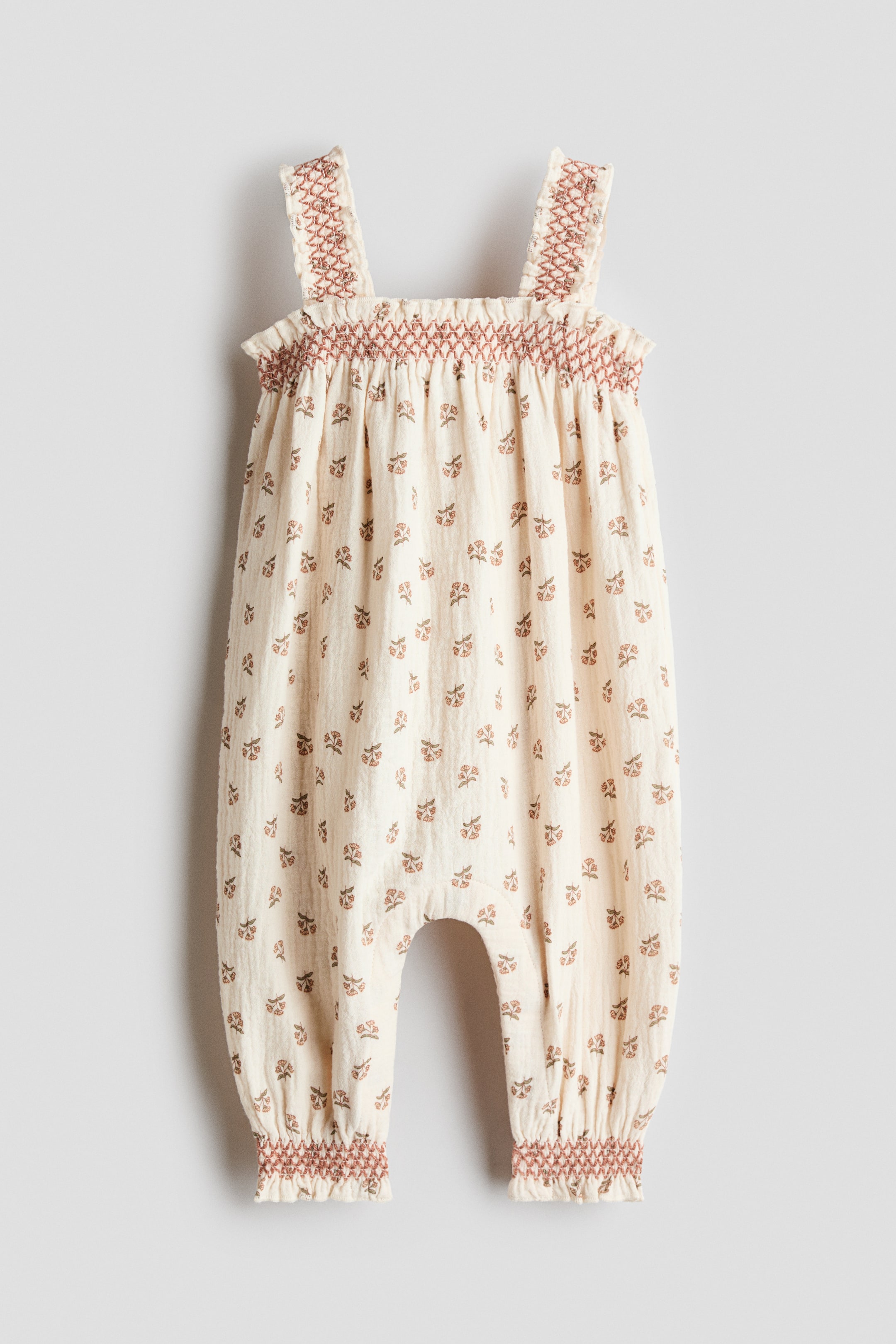 View larger image: A cream baby romper is presented facing front, featuring a smocked bodice and wide smocked shoulder straps. The romper is adorned with a repeating pattern of small brown and red floral motifs and is finished with smocked cuffs at the ankle openings.