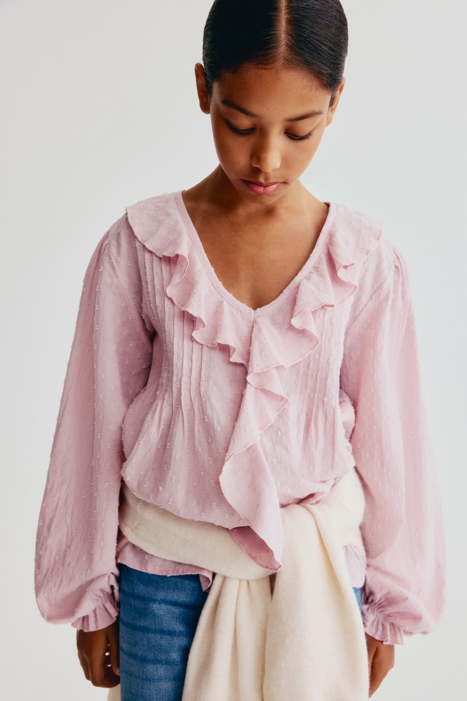 The light pink blouse is showcased in a front-facing view, featuring a textured fabric with small dots, a ruffled V-neckline, and pintucks on the front bodice, along with long sleeves ending in gathered, elasticated cuffs. - 1