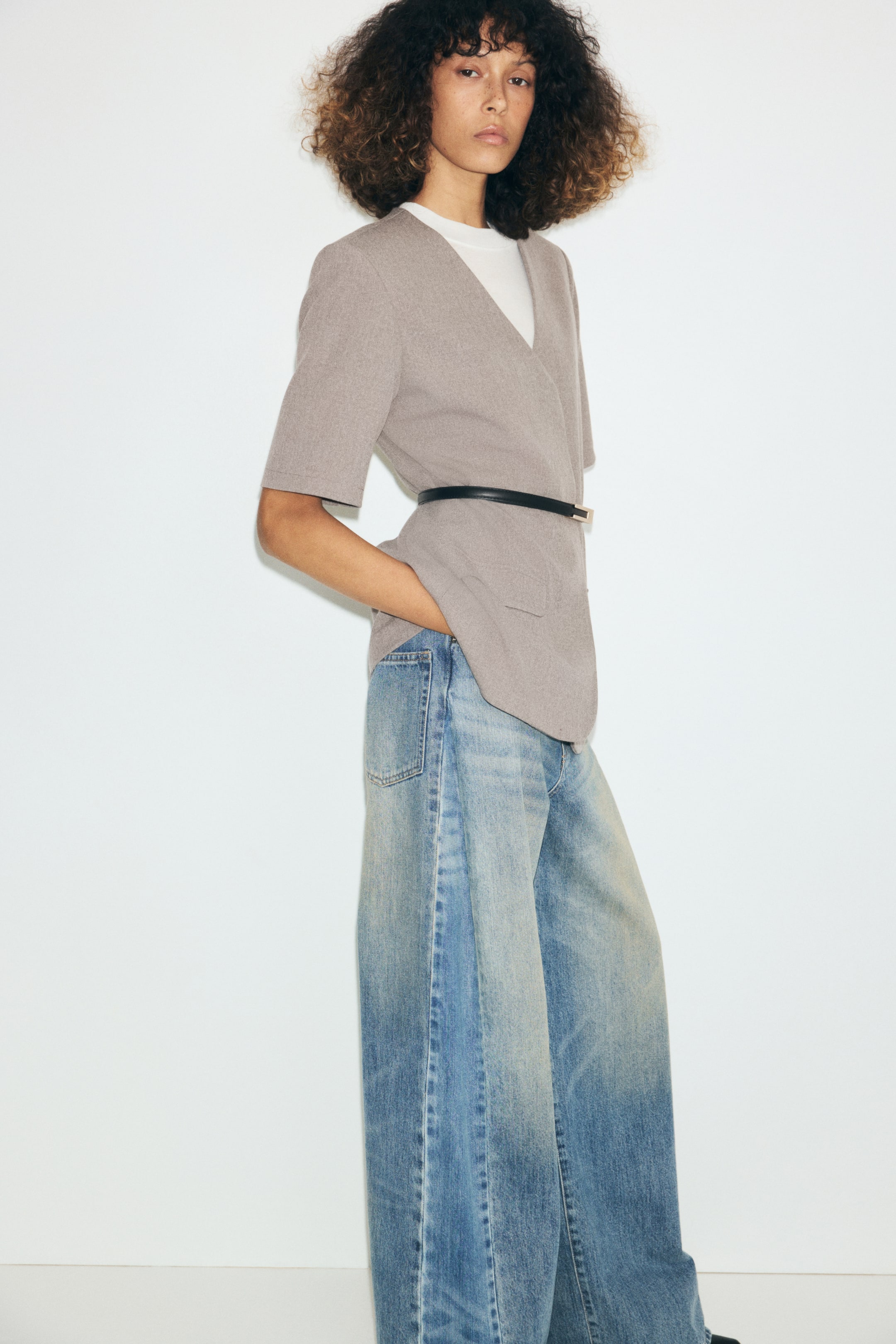 View larger image: A taupe, short-sleeve blazer featuring a lapel-free V-neckline is worn by the model, cinched at the waist with a slim black belt. The blazer is layered over a white T-shirt and paired with wide-leg blue jeans.