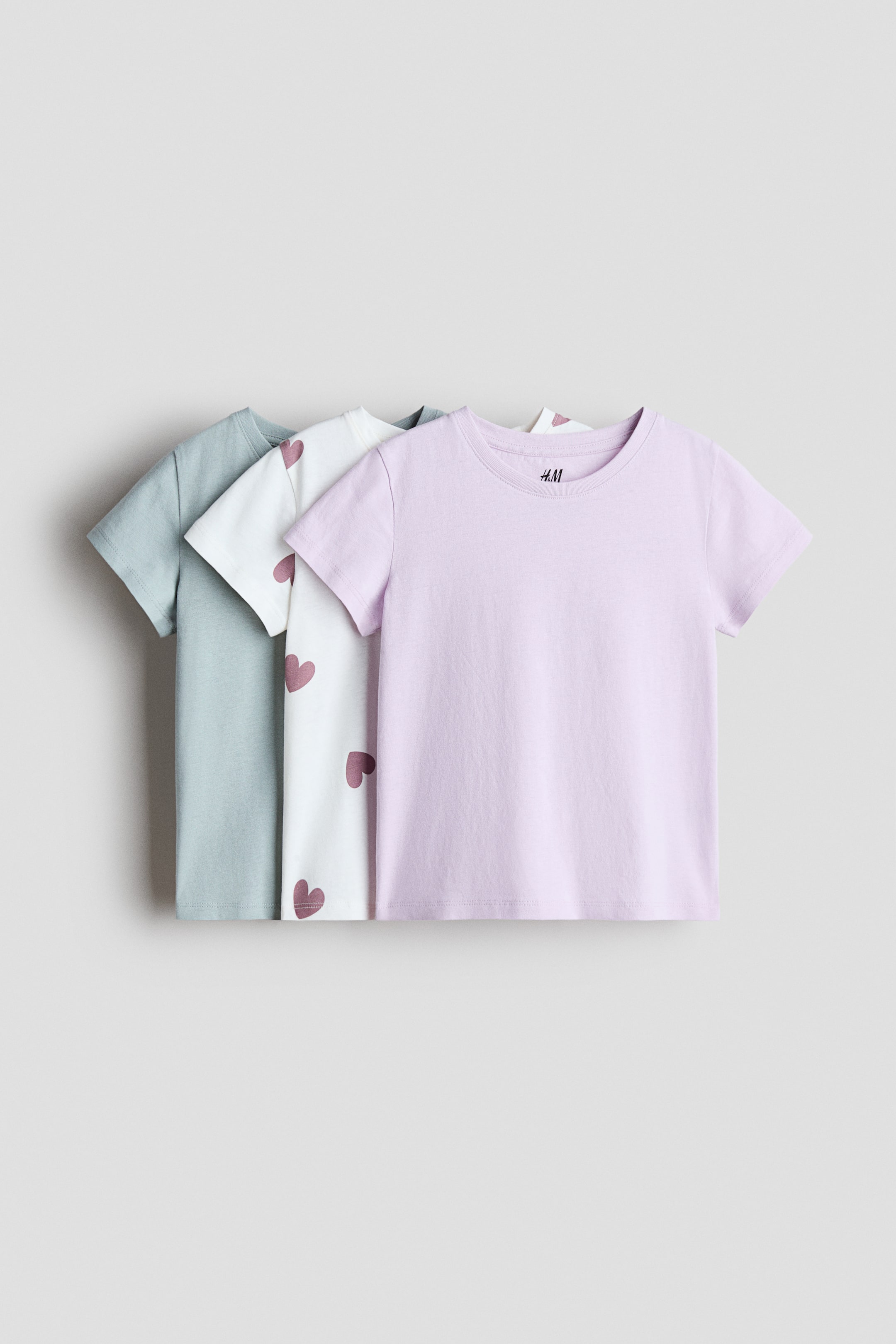 拡大画像を表示: 3枚のTシャツがその前面を見せており、それぞれ丸いネックラインとストレートカットの裾を持っています。前面には淡い紫色の無地のTシャツがあり、その後ろには濃いピンクのハート柄が施された白いTシャツが見え、さらにその後ろには淡い緑色の無地のTシャツが現れています。