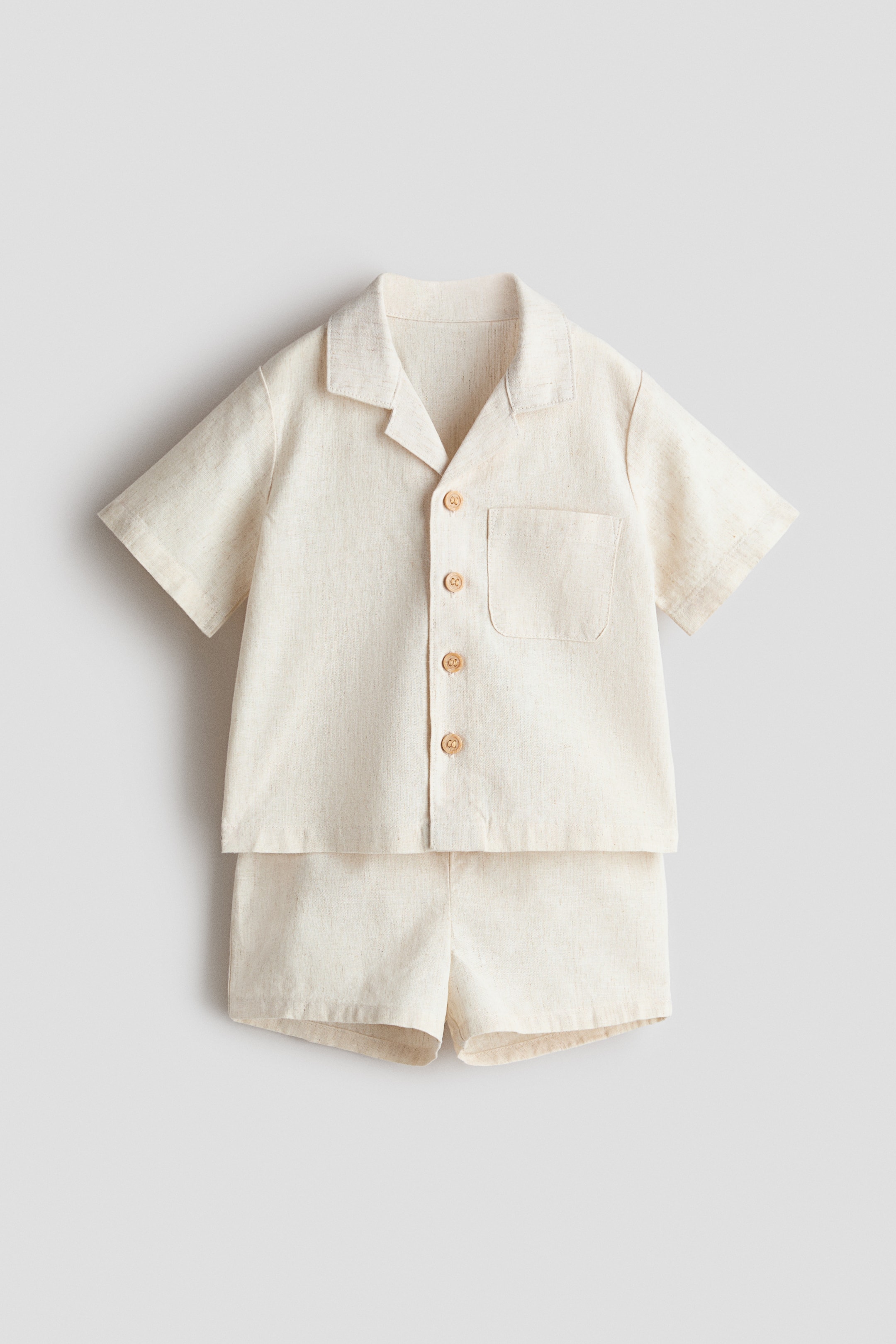 View larger image: A set comprising a short-sleeved shirt and matching shorts in a linen-cotton weave is shown from the front. The shirt features a resort collar, wood-effect buttons down the front, a chest pocket and dropped shoulders, while the shorts have an adjustable, elasticated waistband and sewn-in turn-ups at the hems.