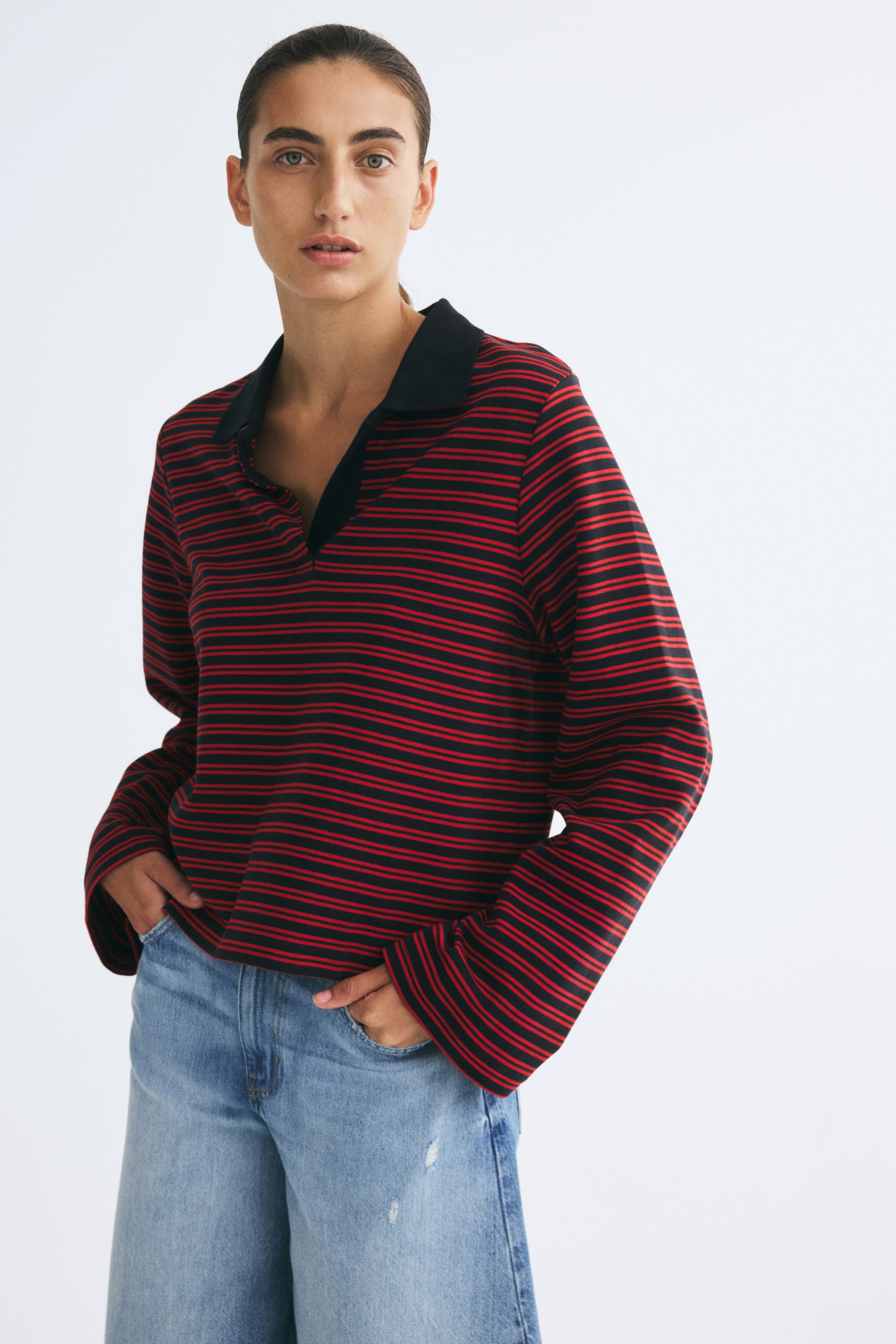 View larger image: The model is wearing a red-and-black striped polo shirt featuring a black open collar, long wide sleeves, and a straight hem. The shirt is styled with light blue denim jeans, and the model’s hands are resting in the front pockets.